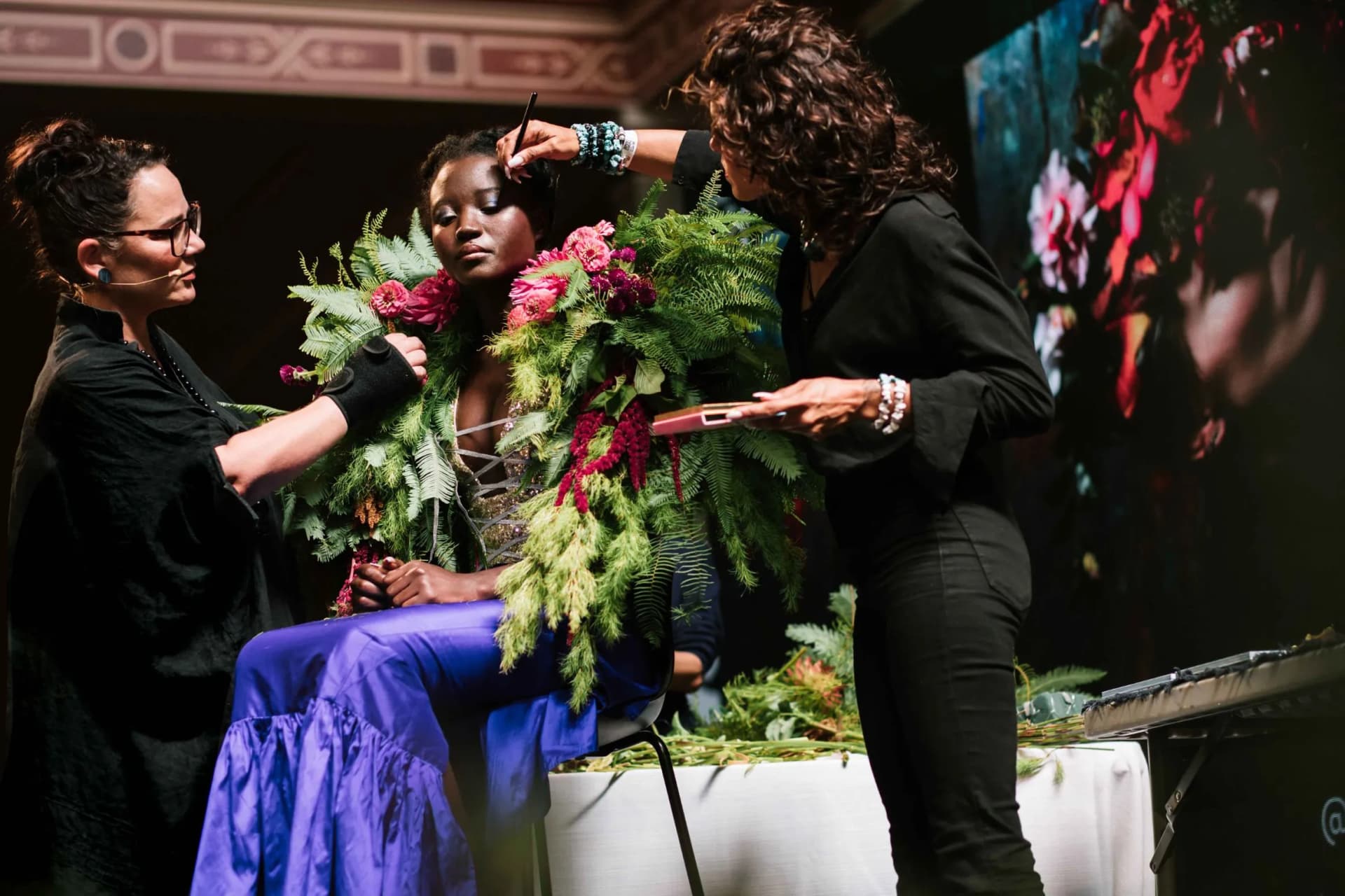 The model is sitting on stage whilst two stylists apply her make-up and drape a sumptuous collar adorned with flowers and ferns around her shoulders.