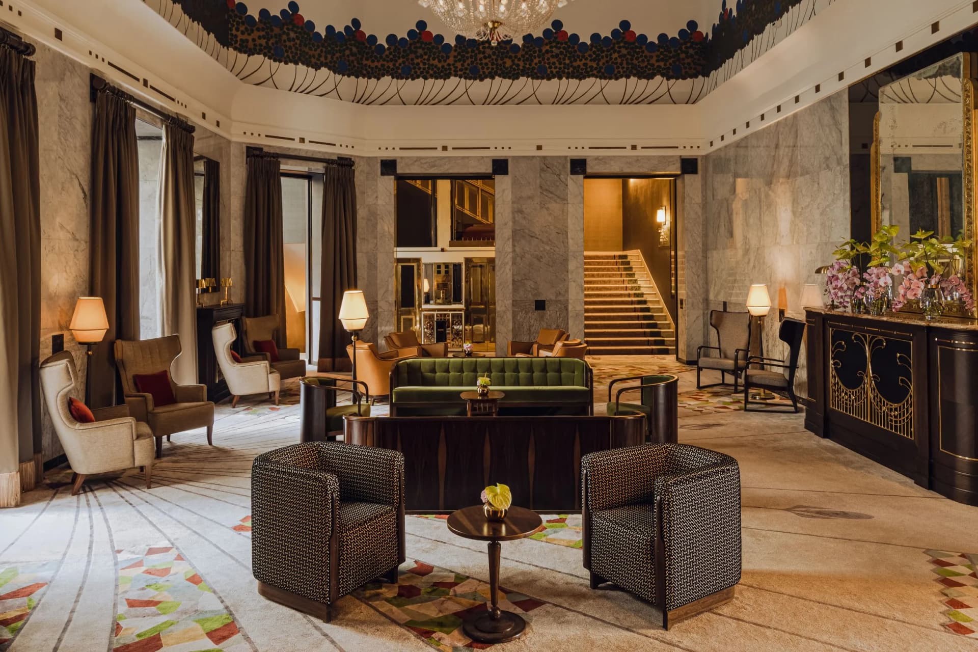 A luxurious marble hotel lobby with comfortable armchairs and a green sofa in the centre, tables with glass tops, gold-plated accents, a magnificent staircase, a chandelier and a floral arrangement.