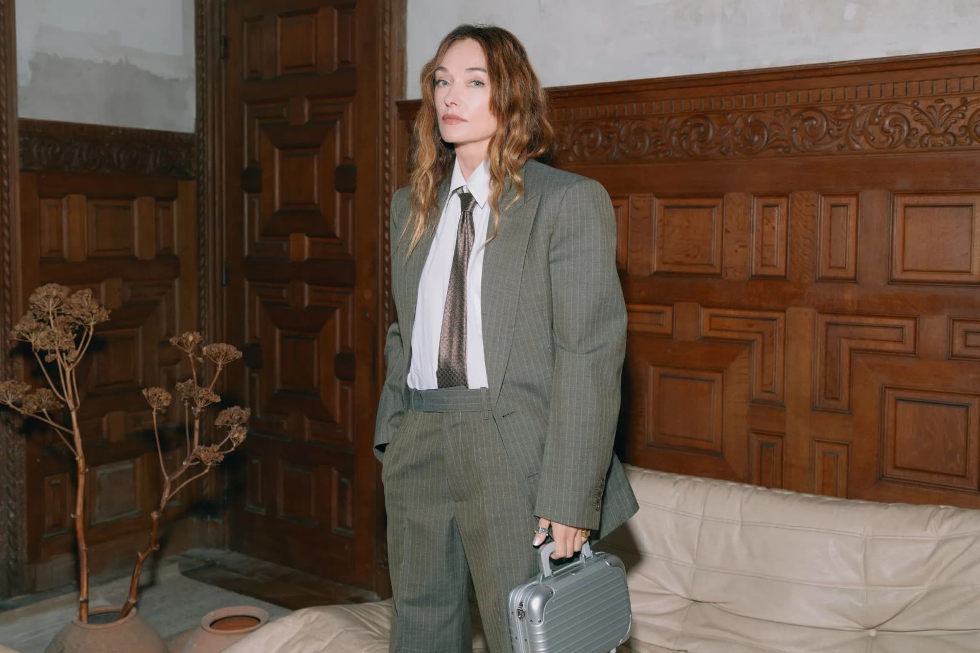 Young woman in a gray suit and tie stands in a wood-paneled room, holding a silver briefcase.