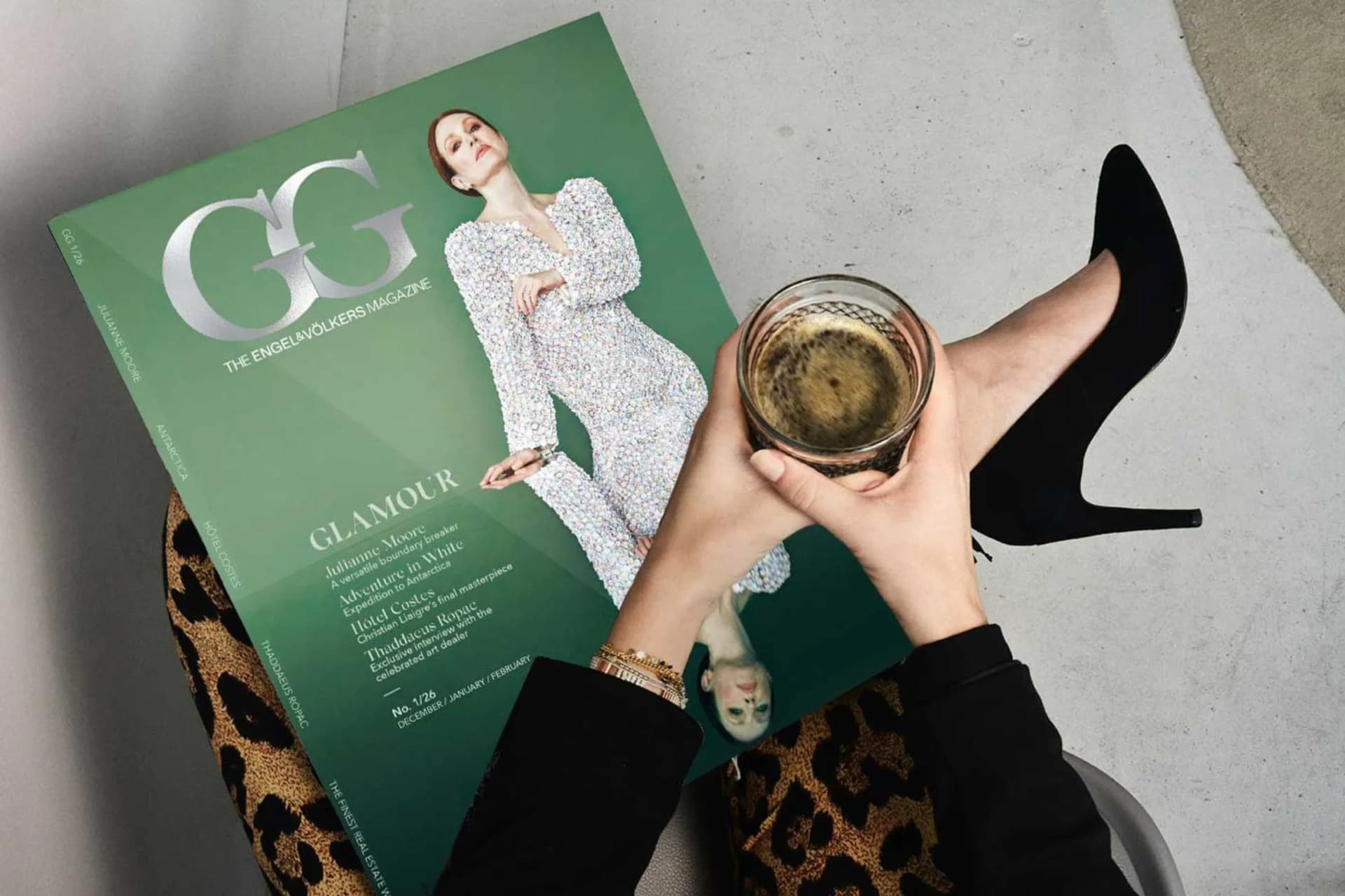 Top-down view of a person in leopard-print pants and black high heel, holding a drink while sitting next to a green fashion magazine.