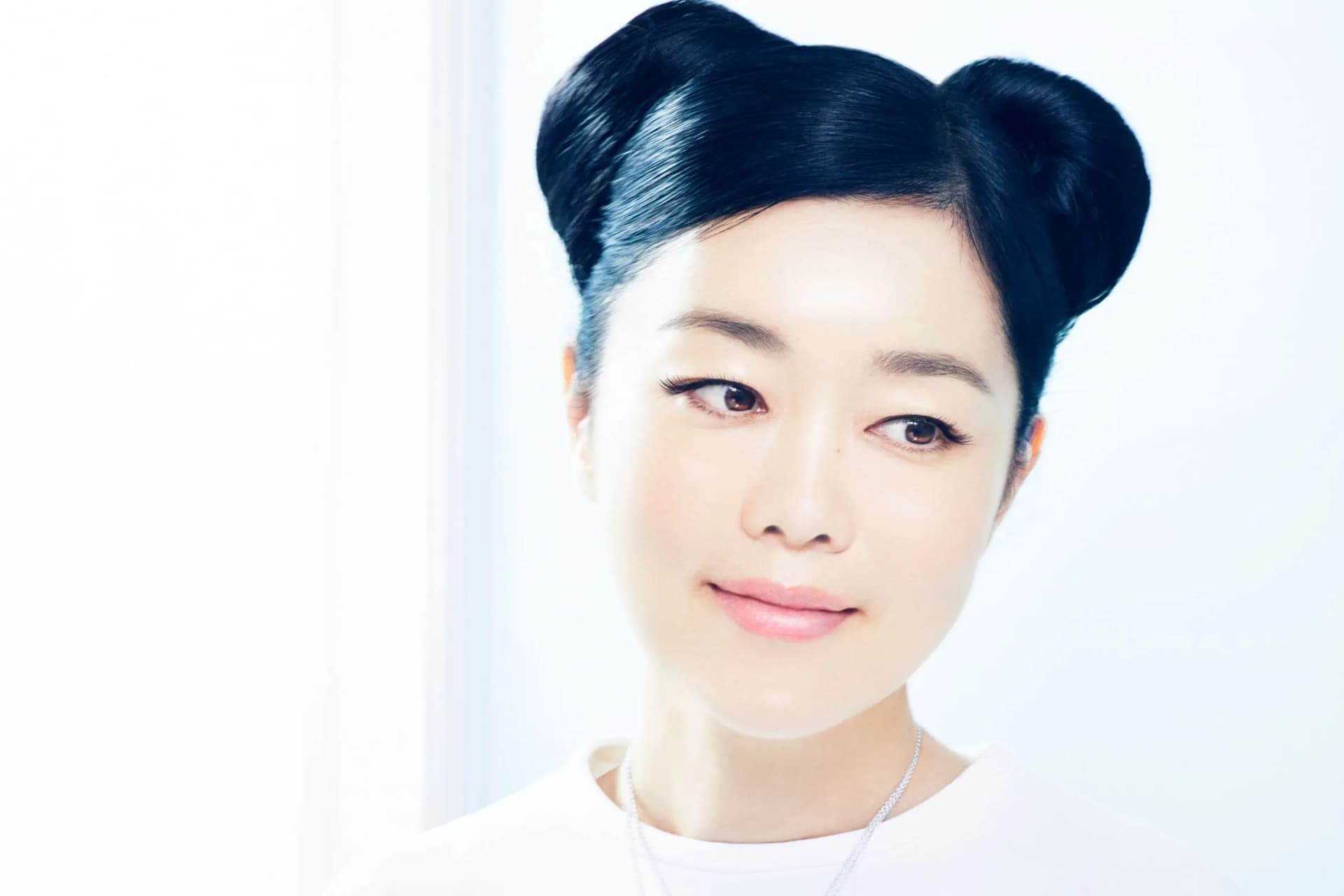 Portrait of an Asian woman with two black buns, soft makeup, gazing to the side in a bright white studio.