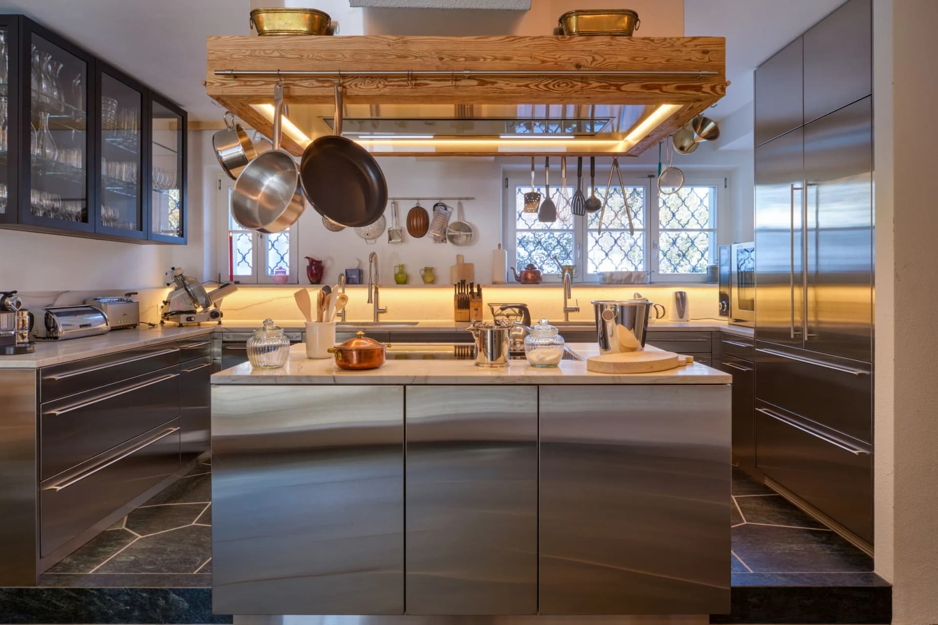 A modern kitchen with a stainless steel kitchen island, pots hanging from a wooden beam, cupboards with glass fronts and warm under-cabinet lighting