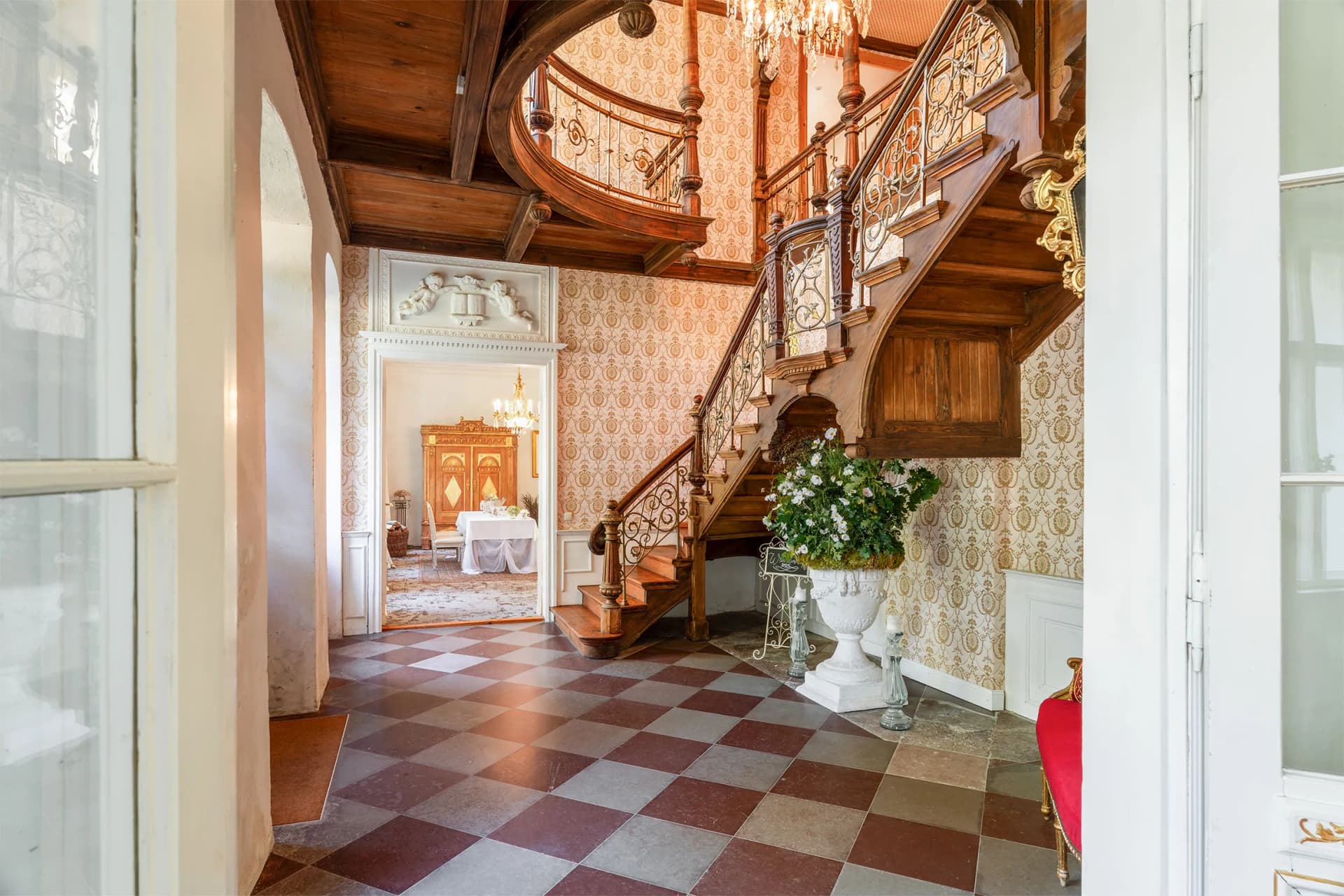 Elegant historic foyer with a curved wooden staircase and ornate iron balustrade, patterned wallpaper, crystal chandelier, and marble pedestal with flowers.