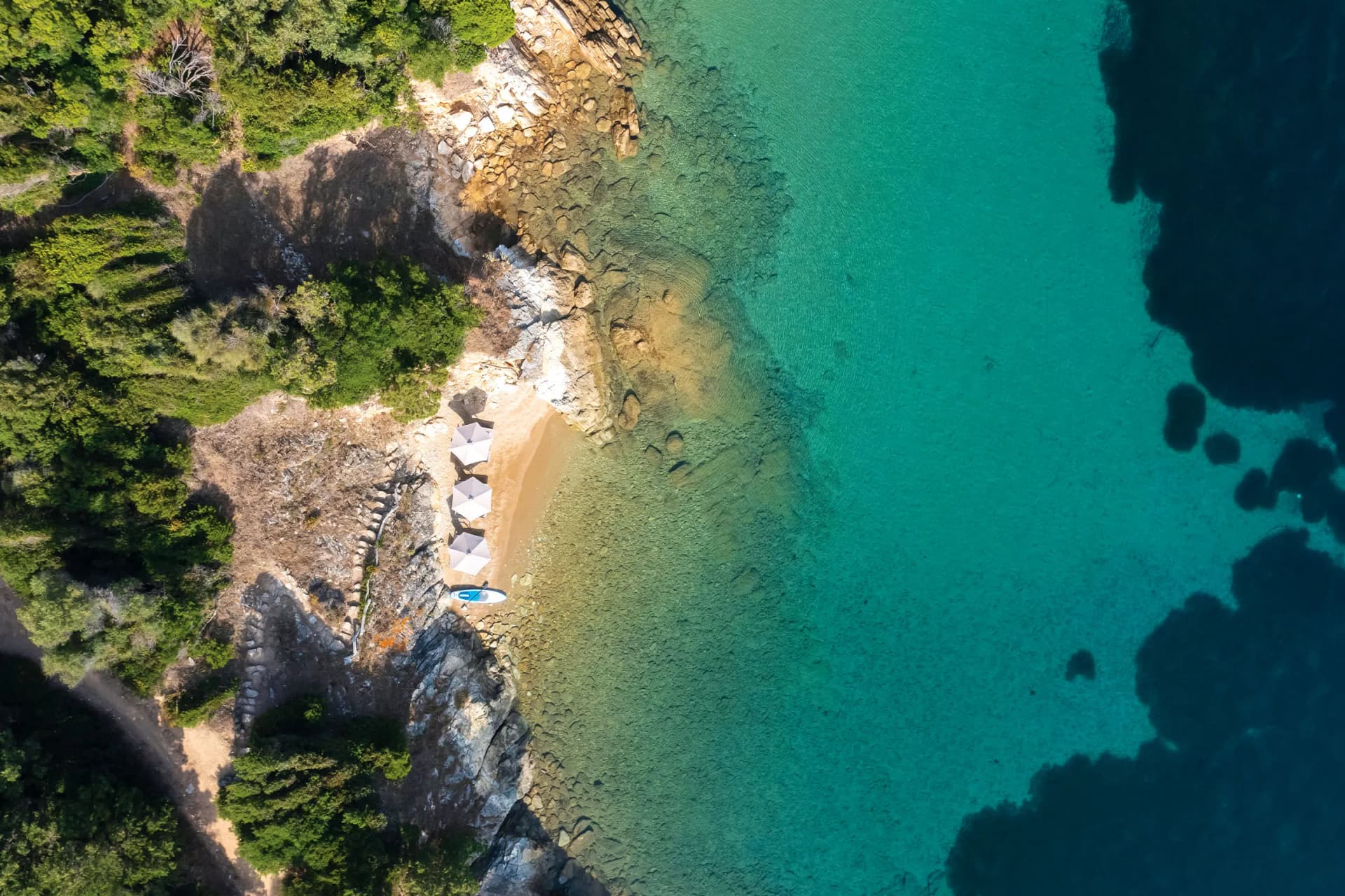 Aerial view of a rocky shoreline with dense trees, turquoise water, and a sandy beach dotted with white umbrellas and paddleboards.