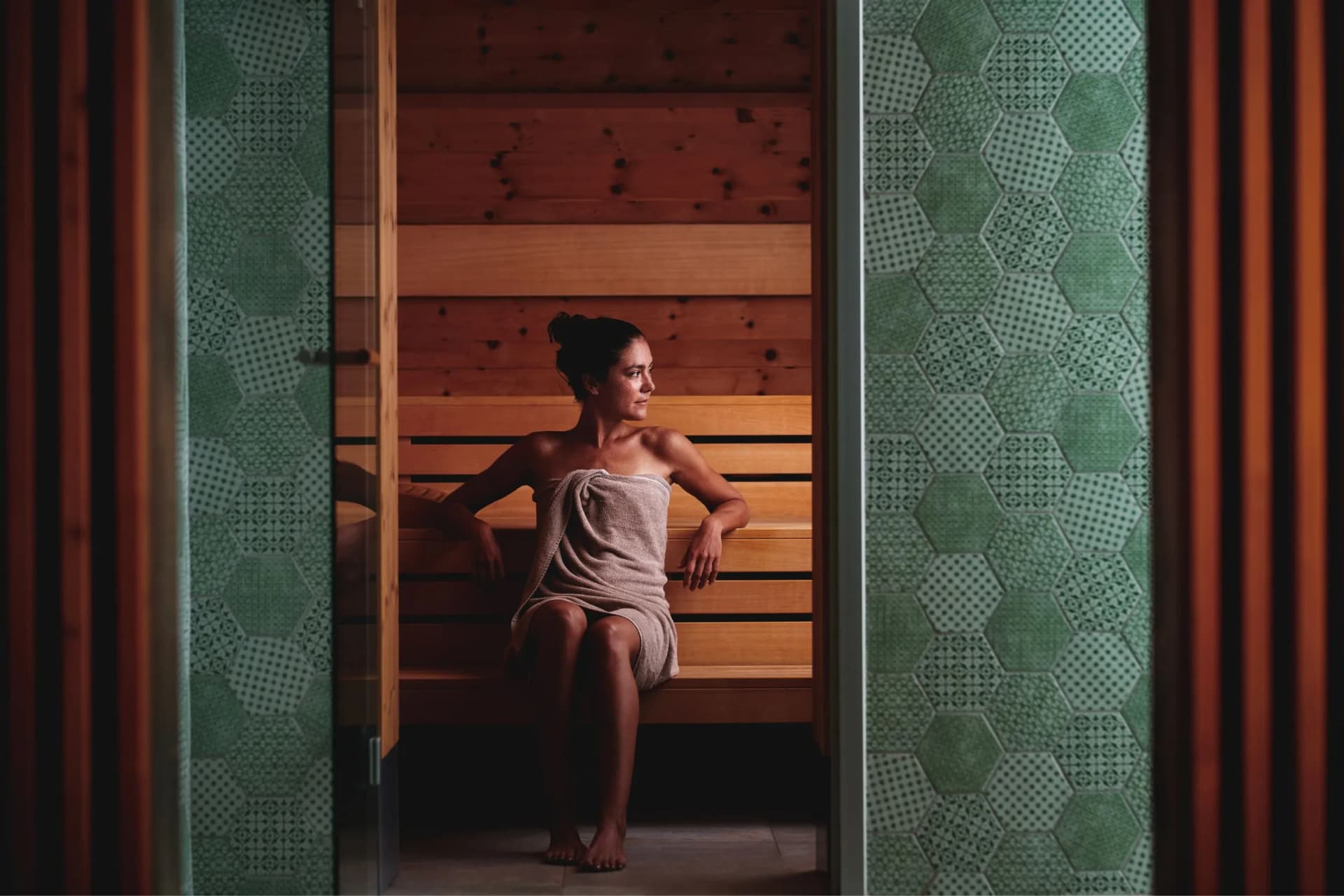 A woman in a towel sits on a wooden bench inside a dimly lit sauna, framed by green hexagonal tiled panels.