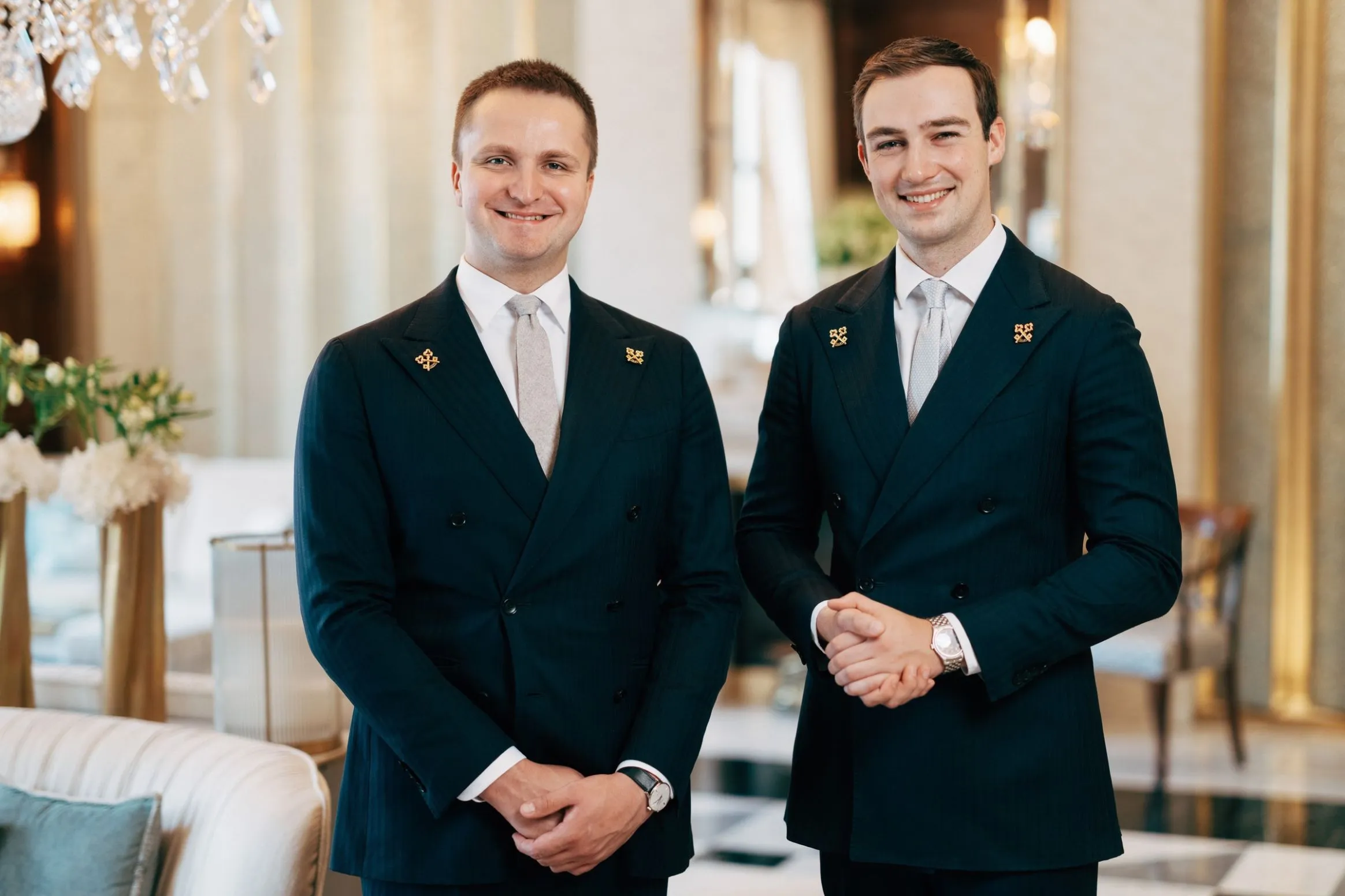 Due uomini, che indossano identici abiti blu navy a doppio petto con cravatte chiare, sorridono nella lussuosa hall di un hotel illuminata da lampadari.