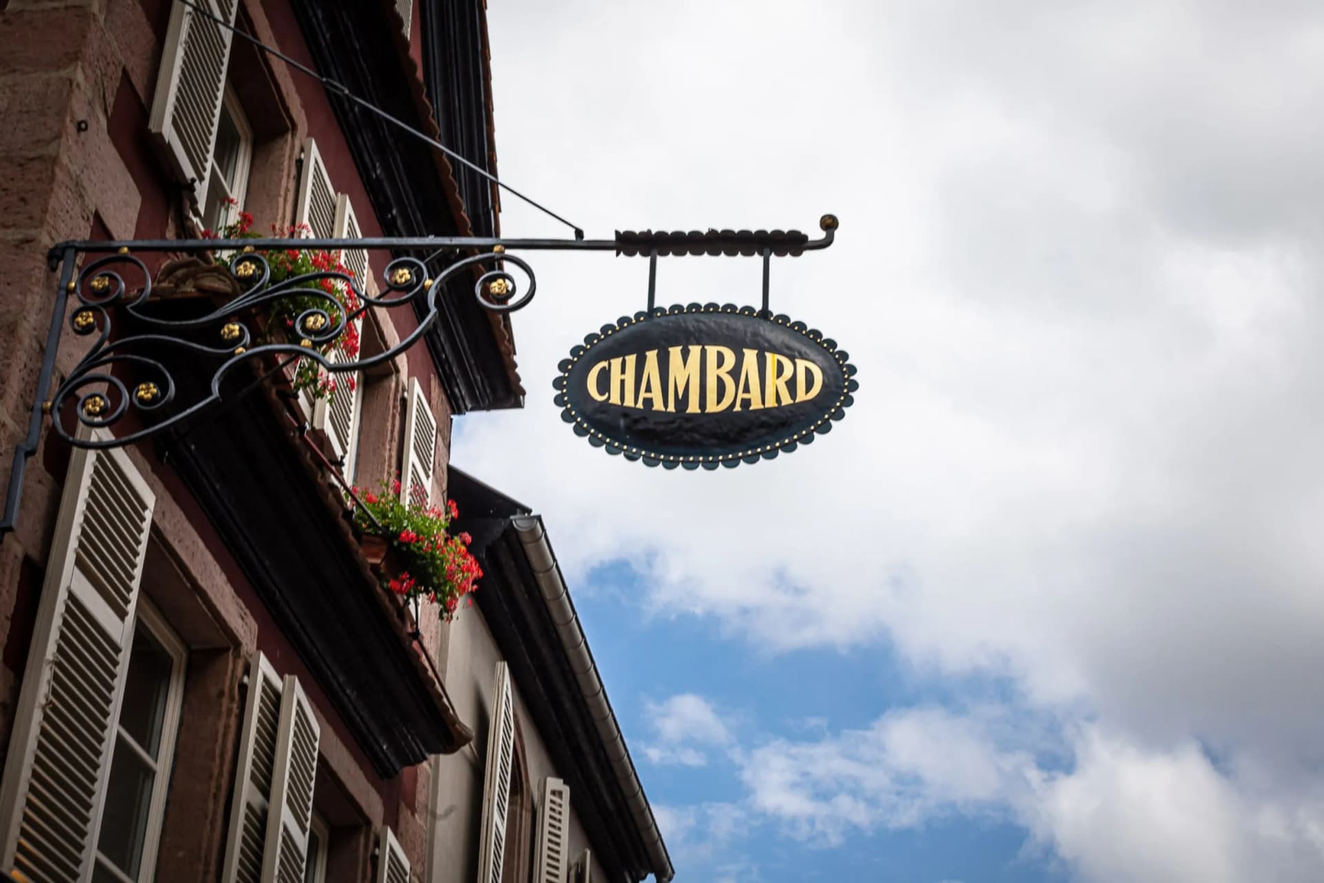 CHAMBARD sign suspended from an ornate wrought-iron bracket on a brick building, with flower boxes and a blue sky.