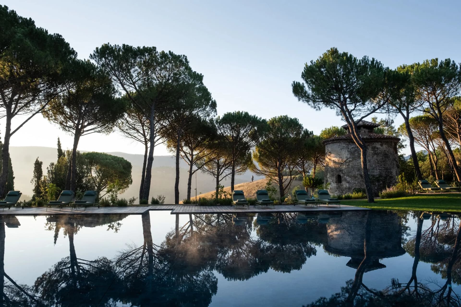 Serene pool reflecting tall pine trees and a cylindrical stone tower, with lounge chairs along the edge beneath a clear sky.