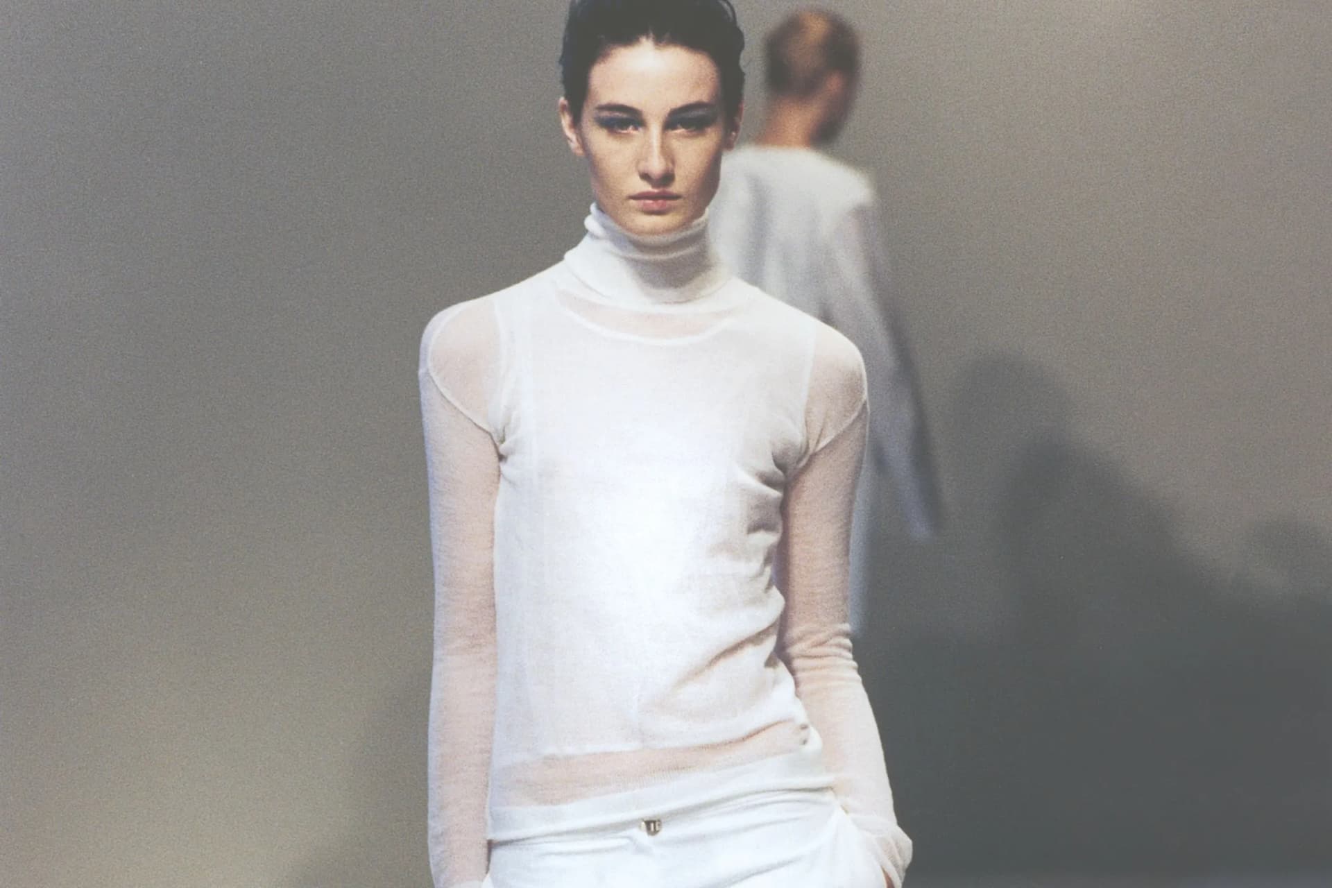 Runway model in a white sheer turtleneck top and white pants, walking toward the camera with a blurred model in the background.