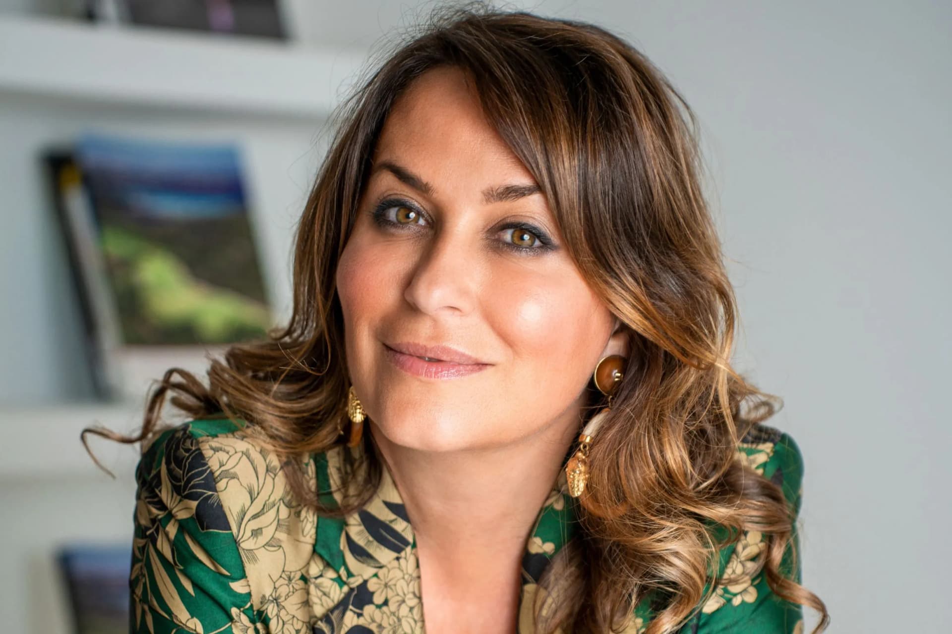 Close-up of a smiling woman with brown hair and hazel eyes, wearing a green floral blouse and gold earrings.