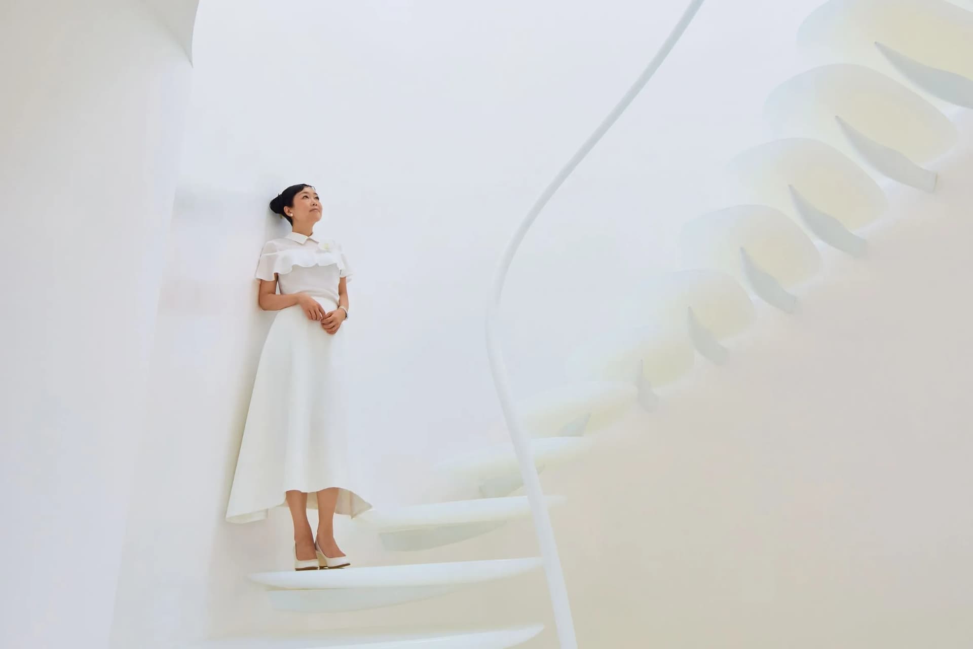 A woman in a white dress stands on a curved white staircase in a bright, minimalist interior.
