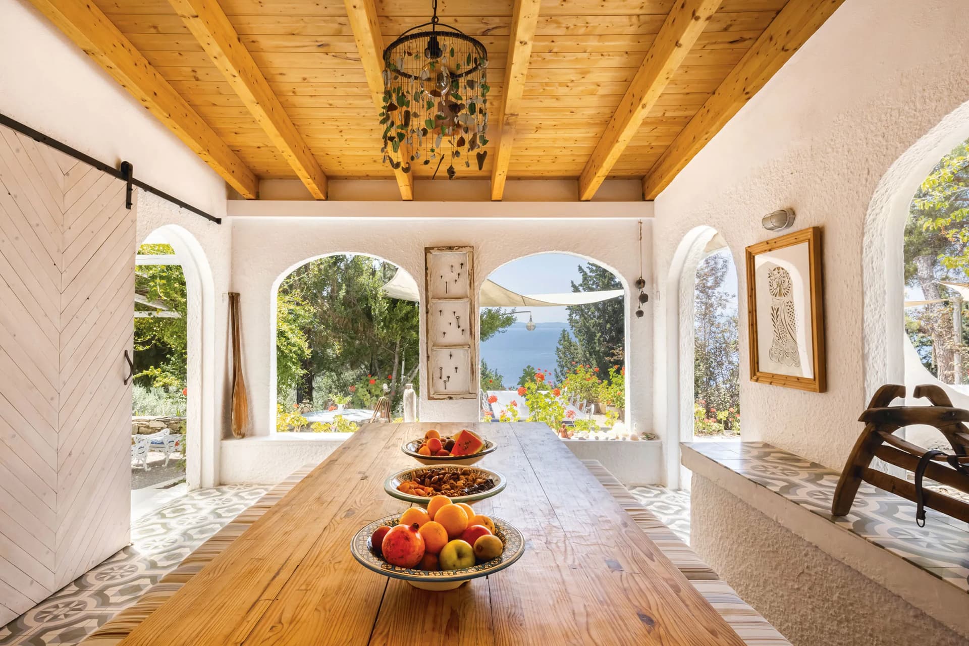 Sunlit covered patio with wooden beams, a long table of fruit bowls, white arches, and a garden and sea view.