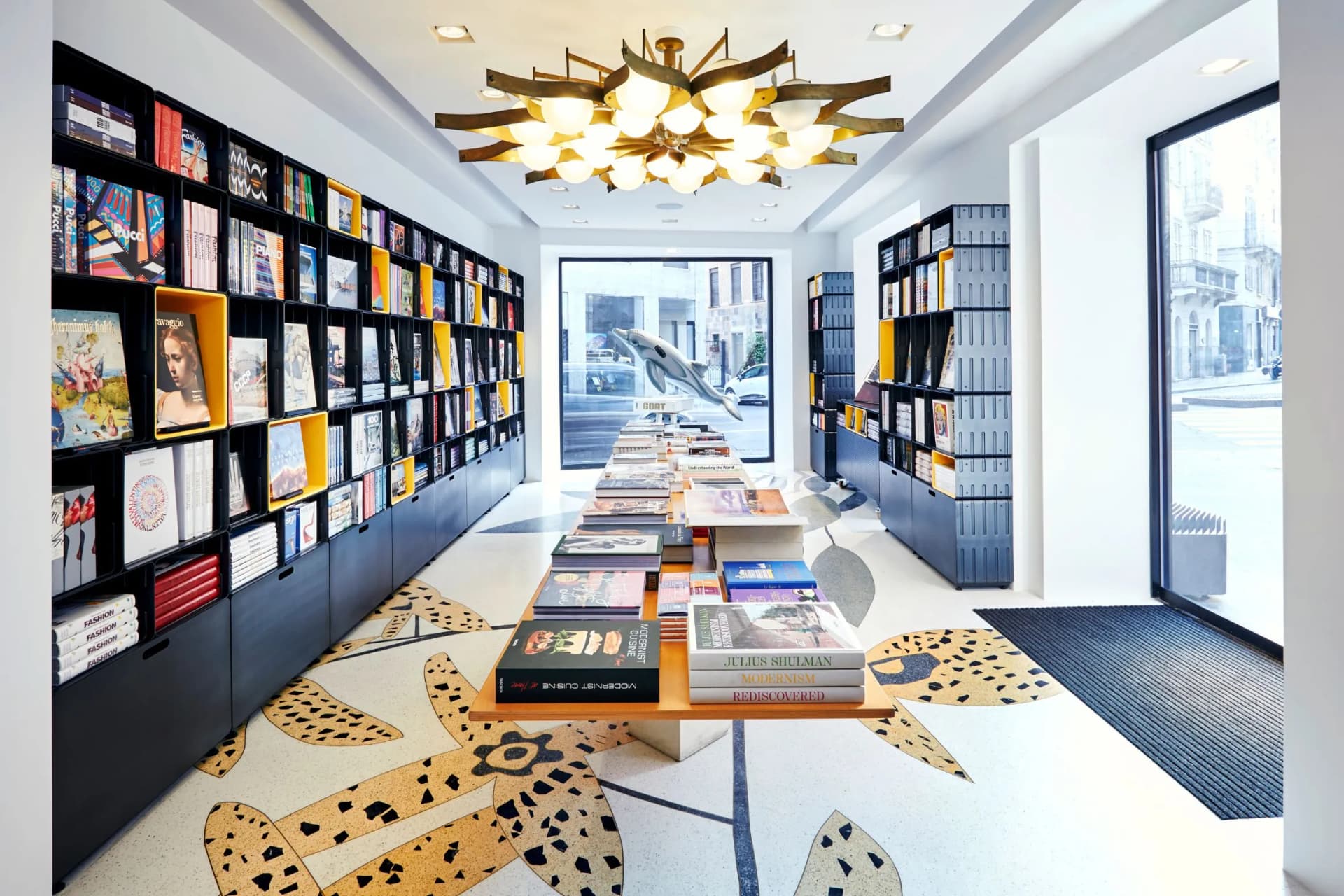 Bright modern bookstore with long central display table, wall-to-wall shelves, a bold patterned floor, and a starburst chandelier.