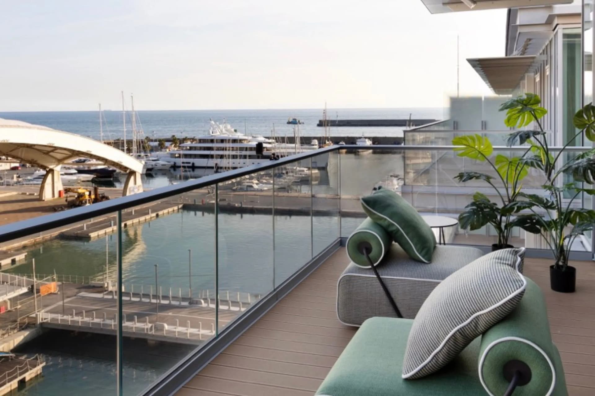 A modern balcony with a glass balustrade and a view of a marina full of yachts; green sun loungers with cushions and potted plants