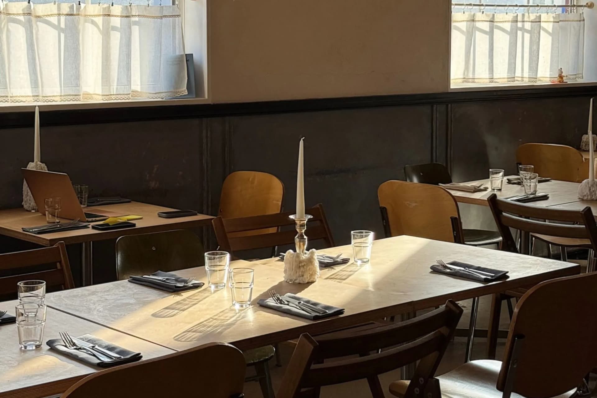 Restaurant dining area with long wooden tables set with napkins, cutlery, glasses, and candles; white lace curtains along the windows.