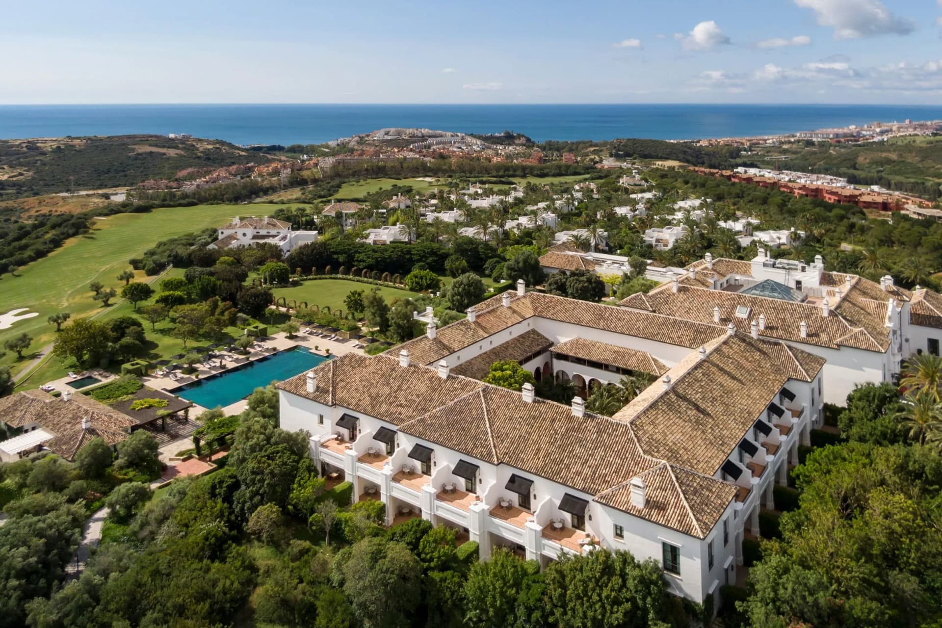 Aerial view of a coastal resort with white villas, terracotta roofs, palm-lined gardens, a pool, and a golf course, overlooking the blue sea.