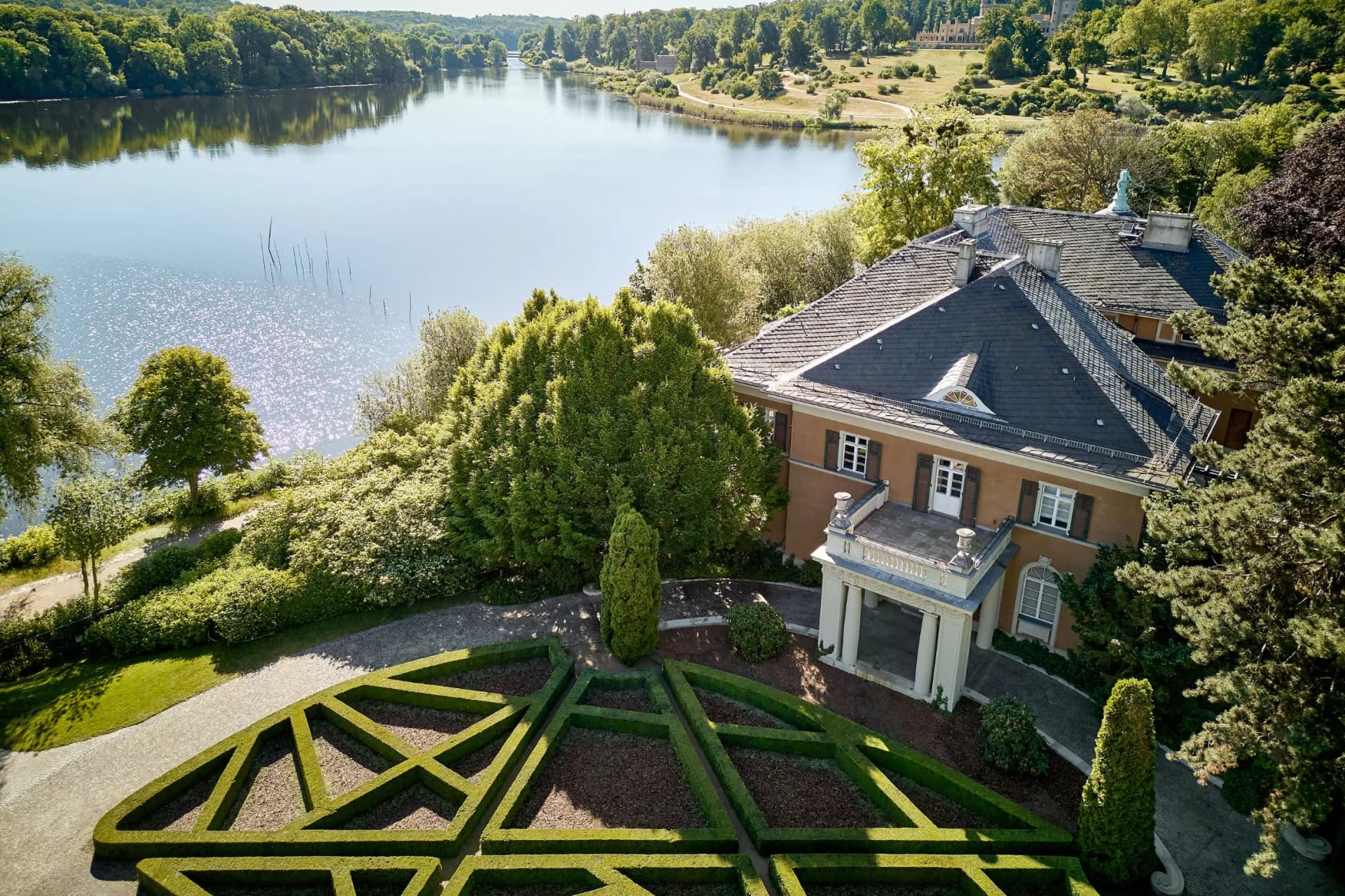 An elegant mansion with a slate roof situated by a tranquil lake, with a geometric maze of green hedges in the foreground and lush trees lining the shore.