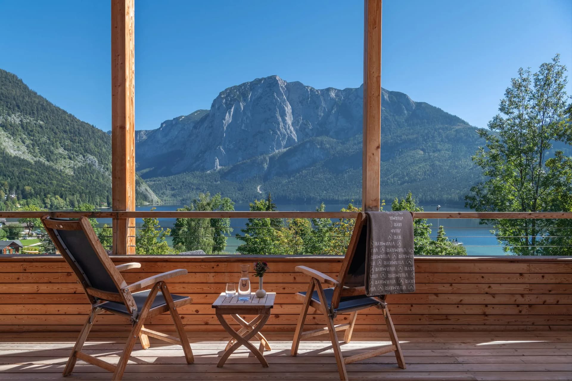 Two wooden lounge chairs on a deck overlooking a mountain lake; a small table between them holds a bottle and two glasses, under a clear blue sky.