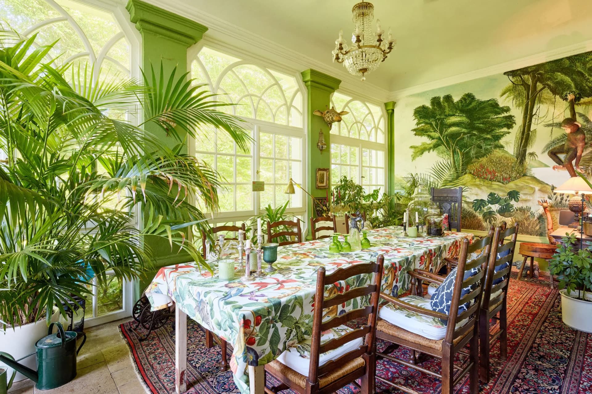 A sun-drenched, tropical-style dining room with large potted palms, white arched windows, green columns, a crystal chandelier and a mural featuring a jungle motif; a long table adorned with flowers, set with glass tableware.