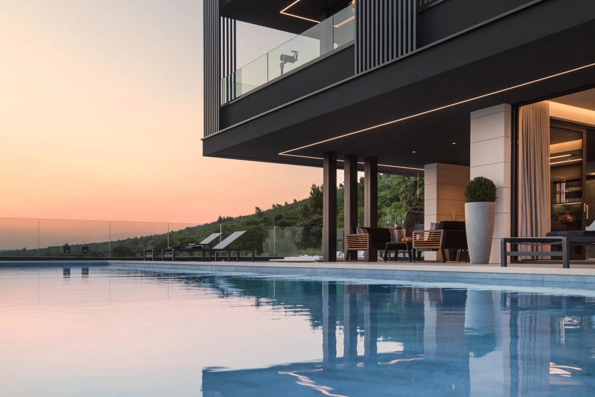 Modern villa with a glass-railed balcony over a reflecting pool at sunset; outdoor seating beneath vertical columns and a tall potted plant.