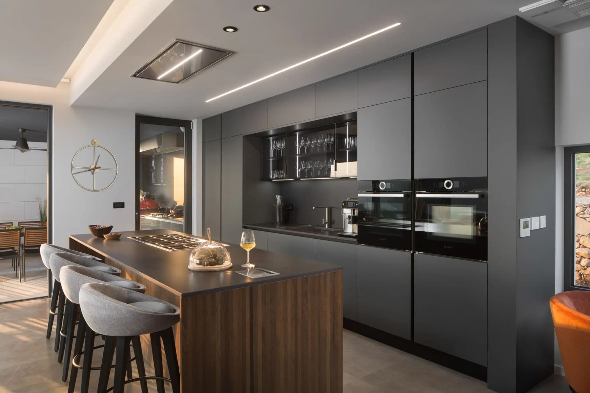 Modern kitchen with dark grey cabinets, built-in ovens, glass-front display of wine glasses, a wooden island with grey stools, and a ceiling light.