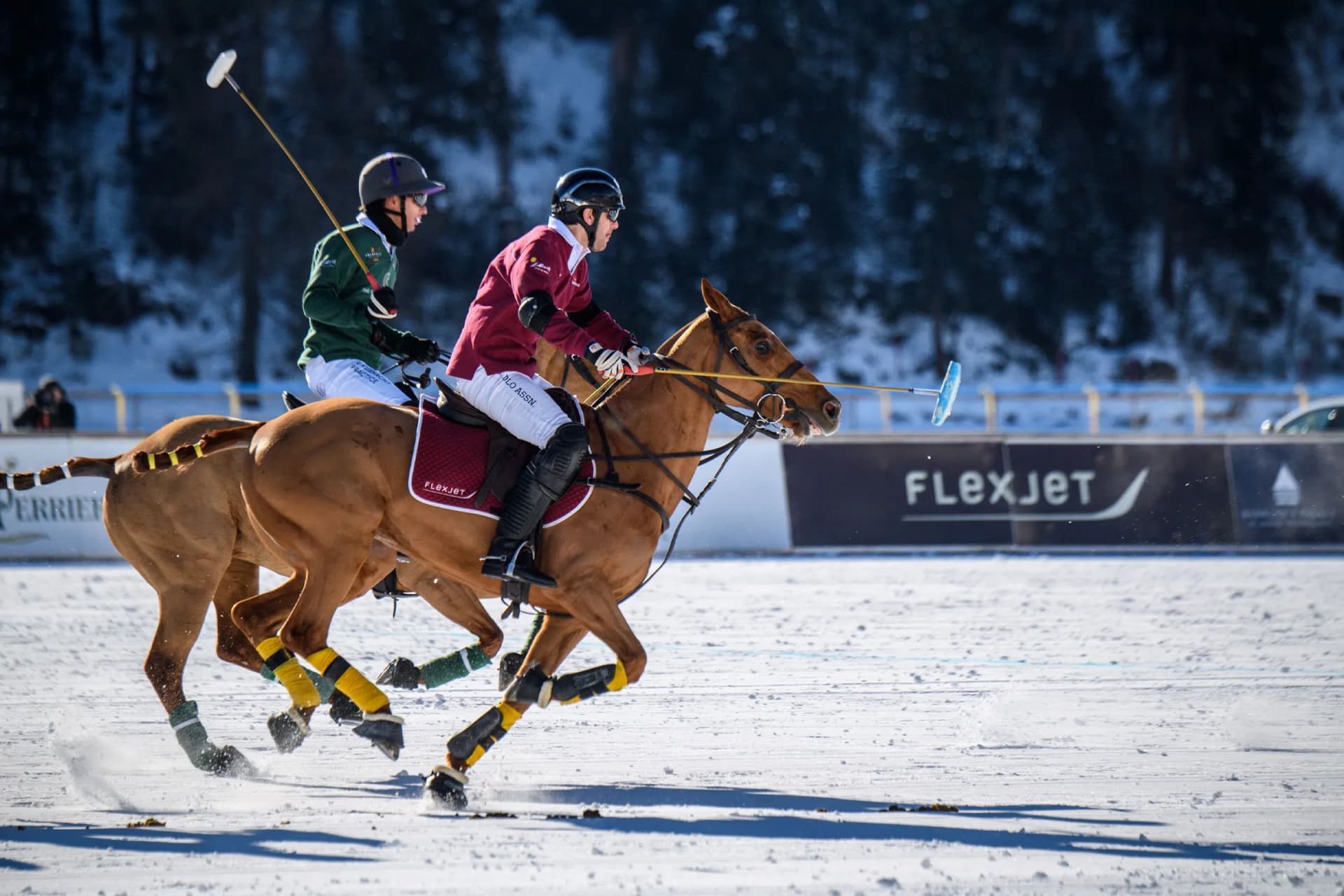 Two riders on horses compete in a snow polo match, swinging mallets on a snow-covered field with banners and a forest backdrop.