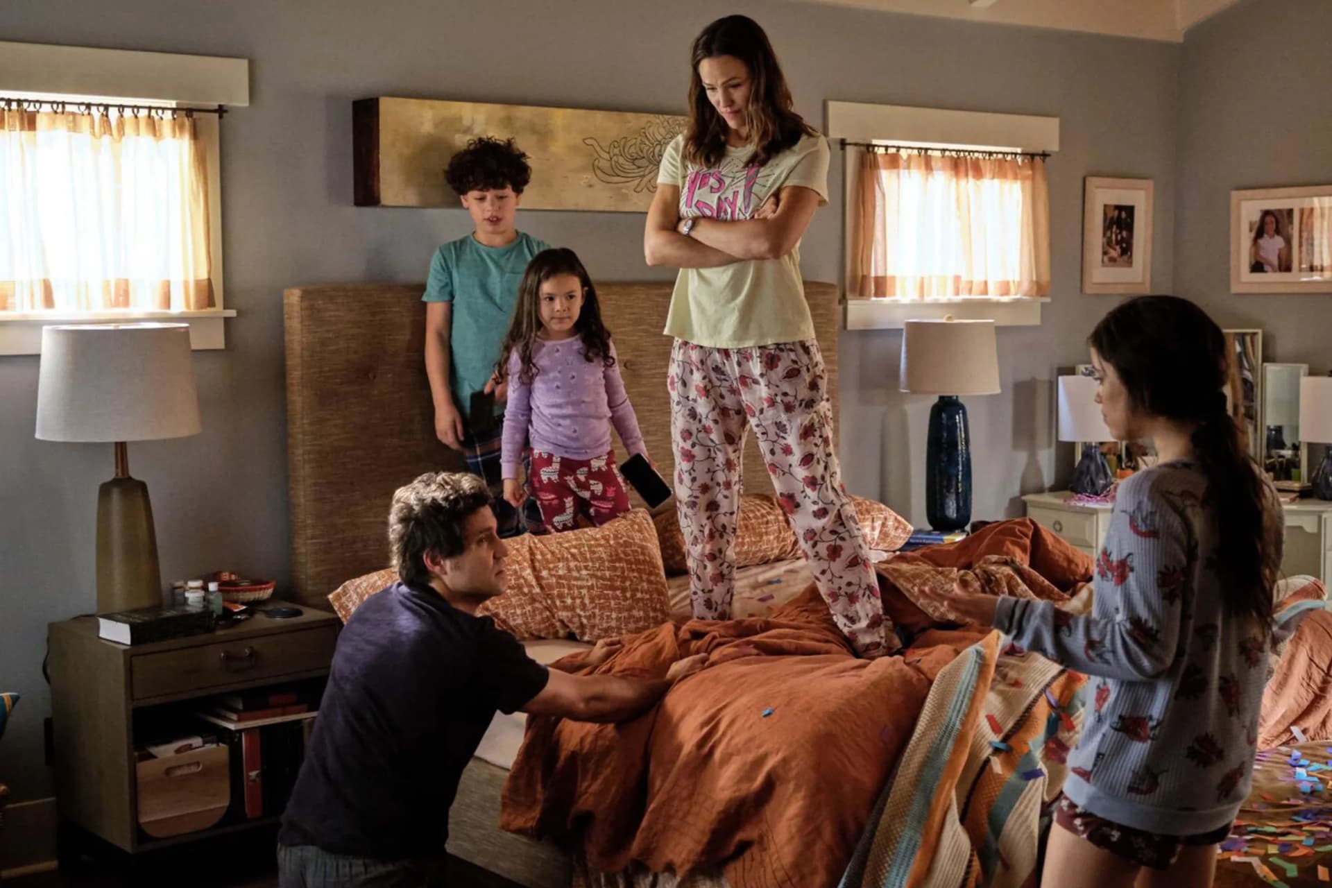 A family in a cozy bedroom; a girl stands on a bed while others watch, as a man crouches by the edge to help her down.