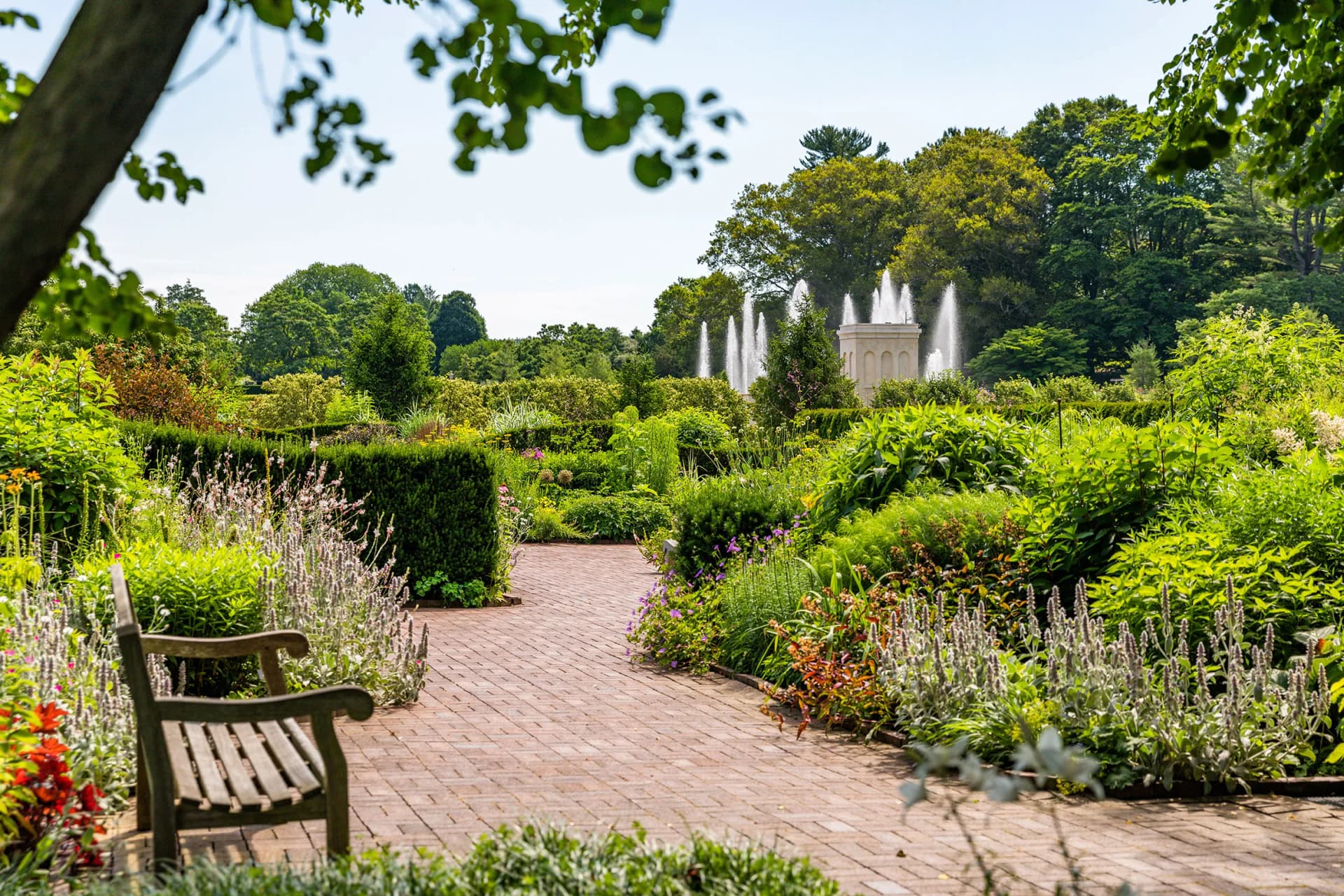 Brick garden path with a wooden bench in the foreground, vibrant flower beds and hedges, and a distant fountain spraying water among trees.