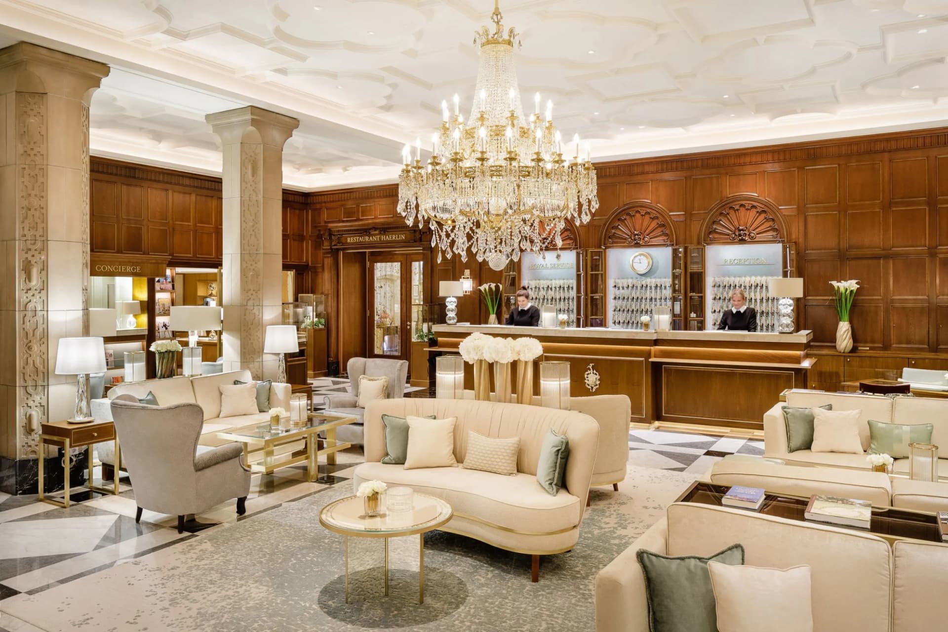 La lussuosa hall di un hotel, con un magnifico lampadario di cristallo, pareti rivestite in legno, pavimenti in marmo, divani color crema e una reception presidiata.