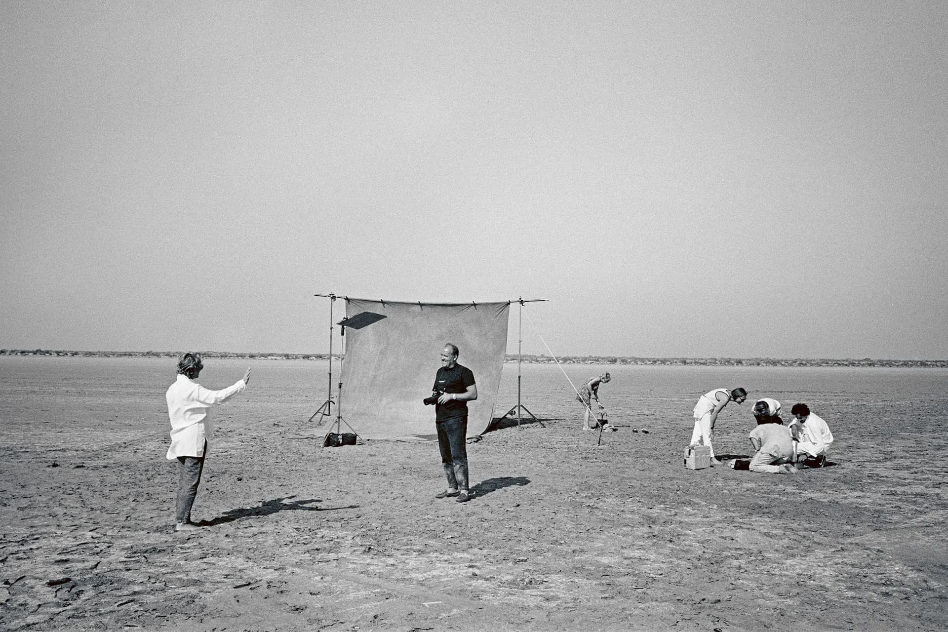 A film crew shoots a scene on a barren, flat desert; a portable backdrop stands behind a man at center while others adjust gear nearby.