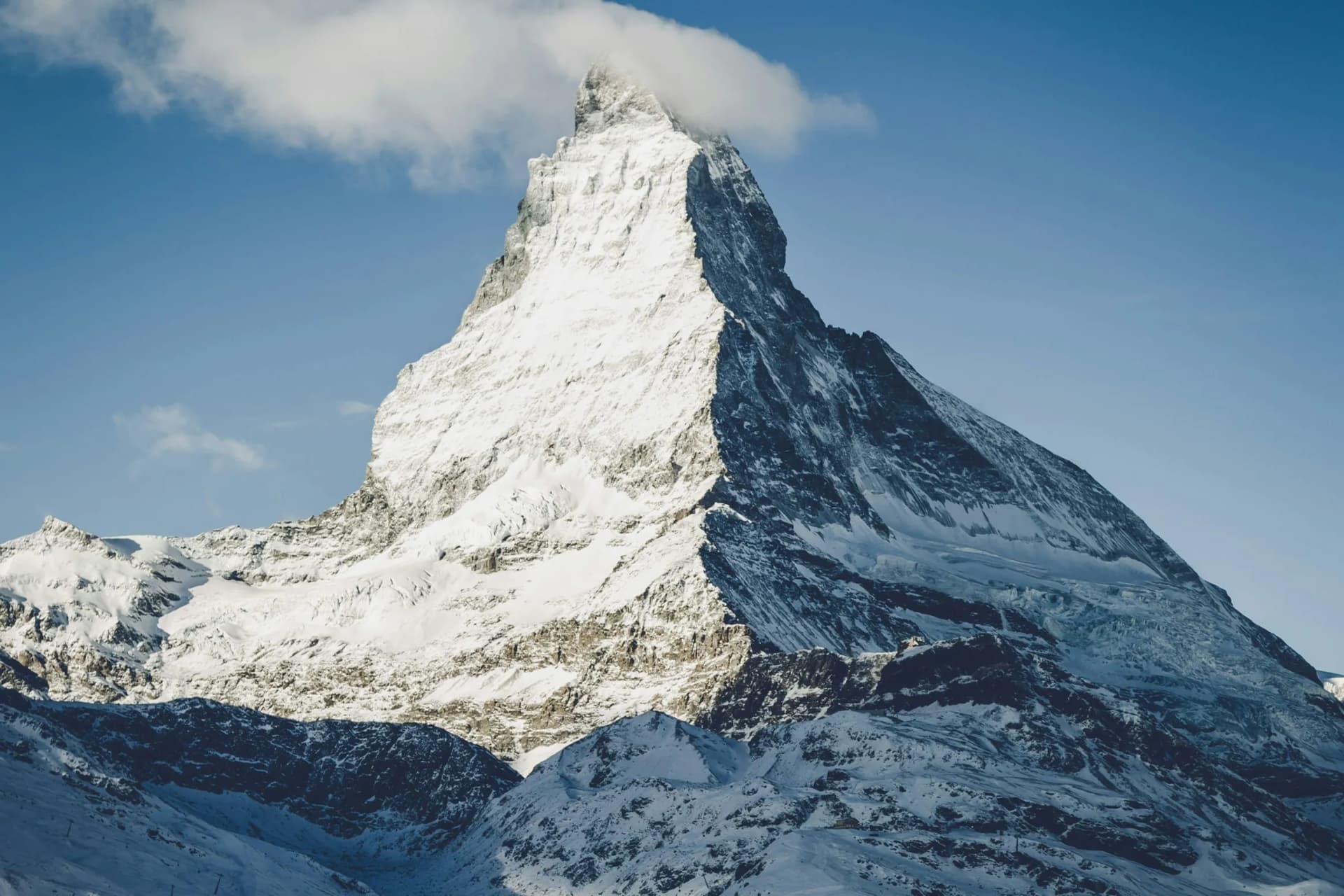 Snow-covered, jagged mountain peak rising above snowfields, with a wispy cloud near the summit under a clear blue sky.