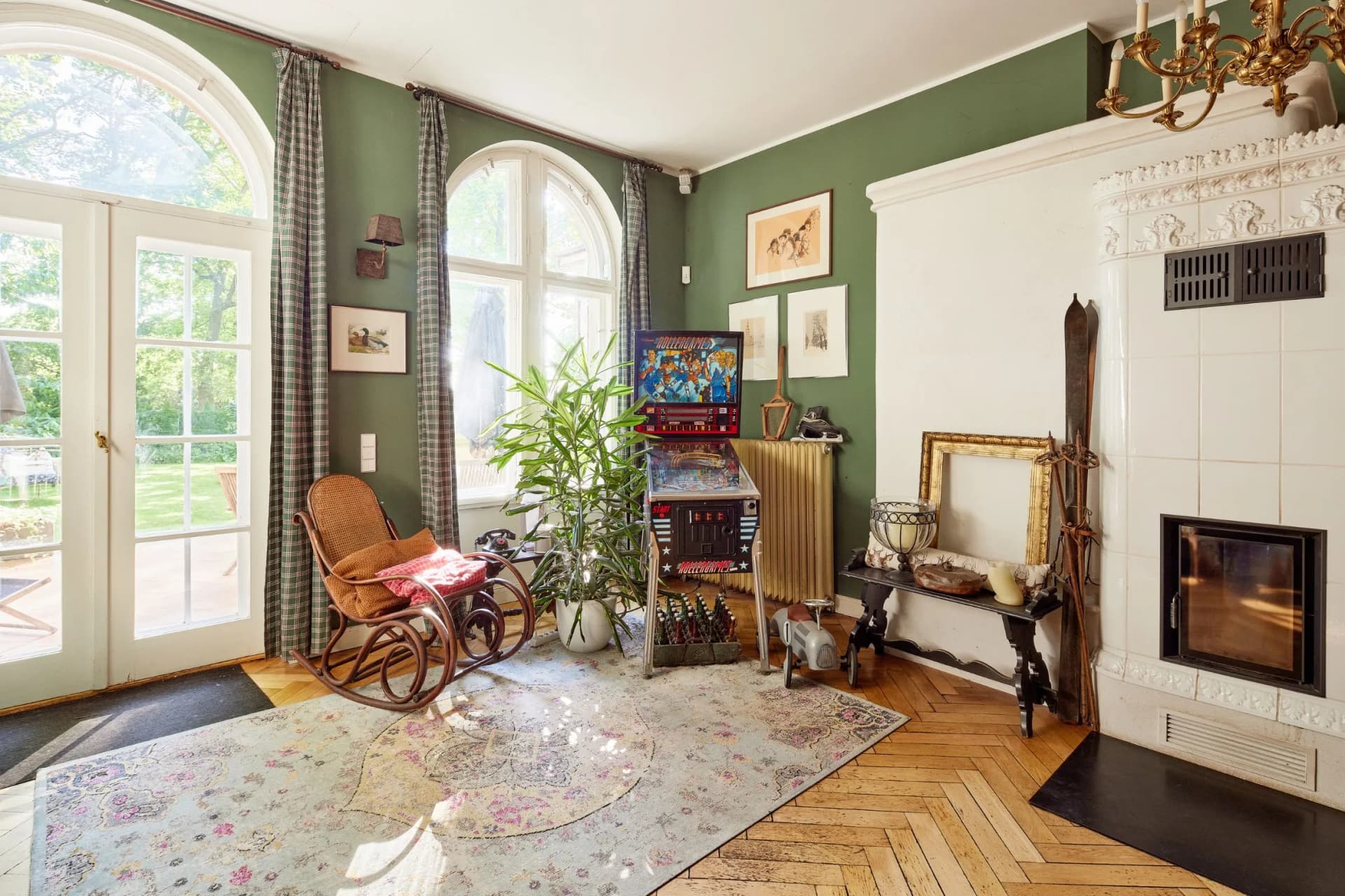 A cosy, vintage-style living room with green walls, arched windows, checked curtains, wicker chairs, a pinball machine, a plant and an ornate white fireplace.