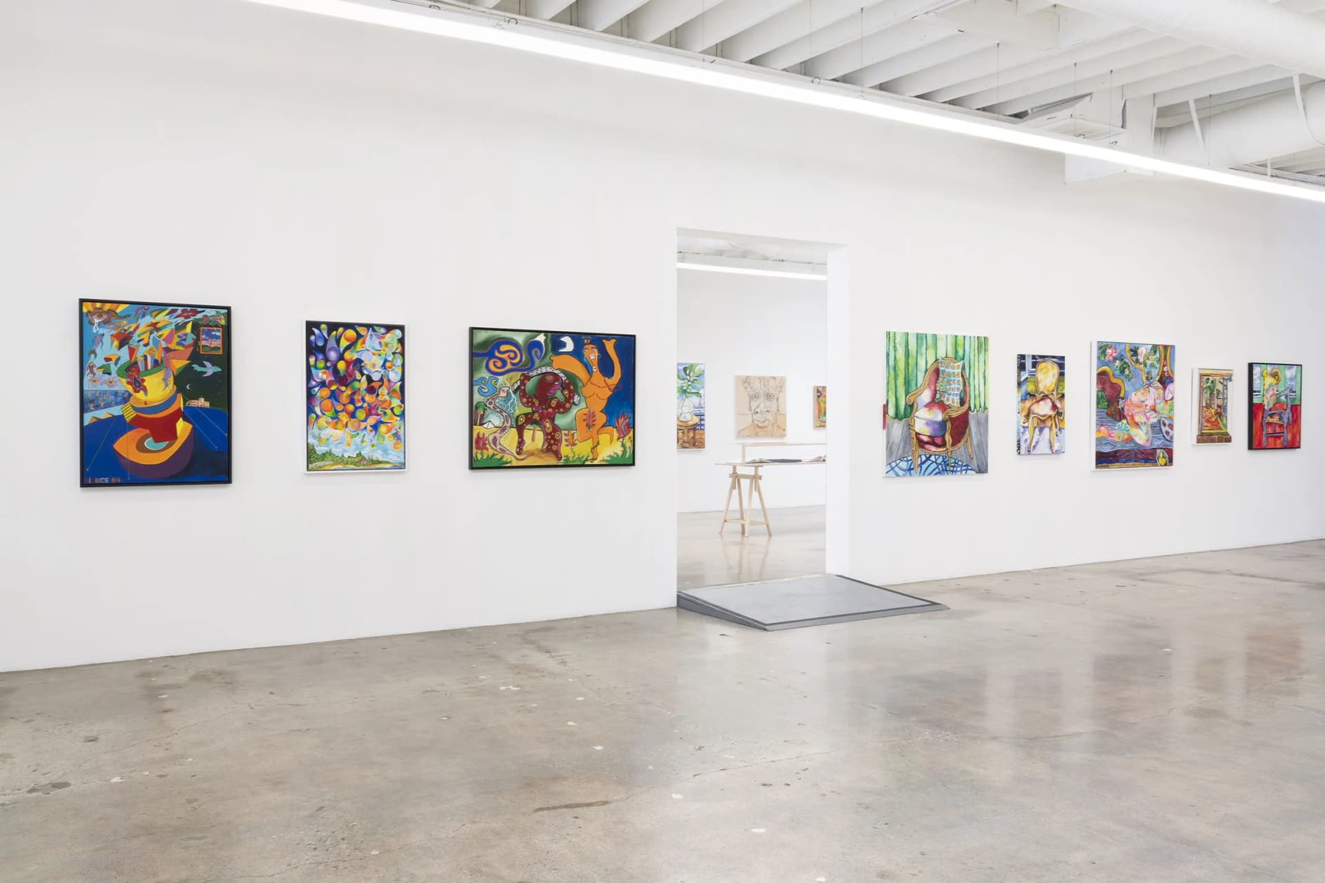 A bright white gallery space with colorful paintings lining the walls and an open doorway to another room with more art.