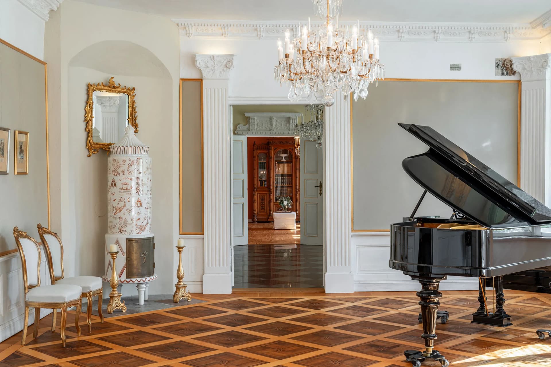 Elegant room with a black grand piano, crystal chandelier, gold-framed mirrors, ornate columns, and a tall porcelain vase beside gilded chairs.