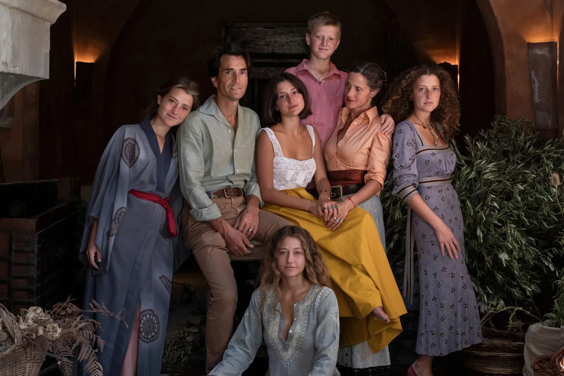 Seven people in vintage clothing pose together in a rustic archway, surrounded by baskets and greenery.