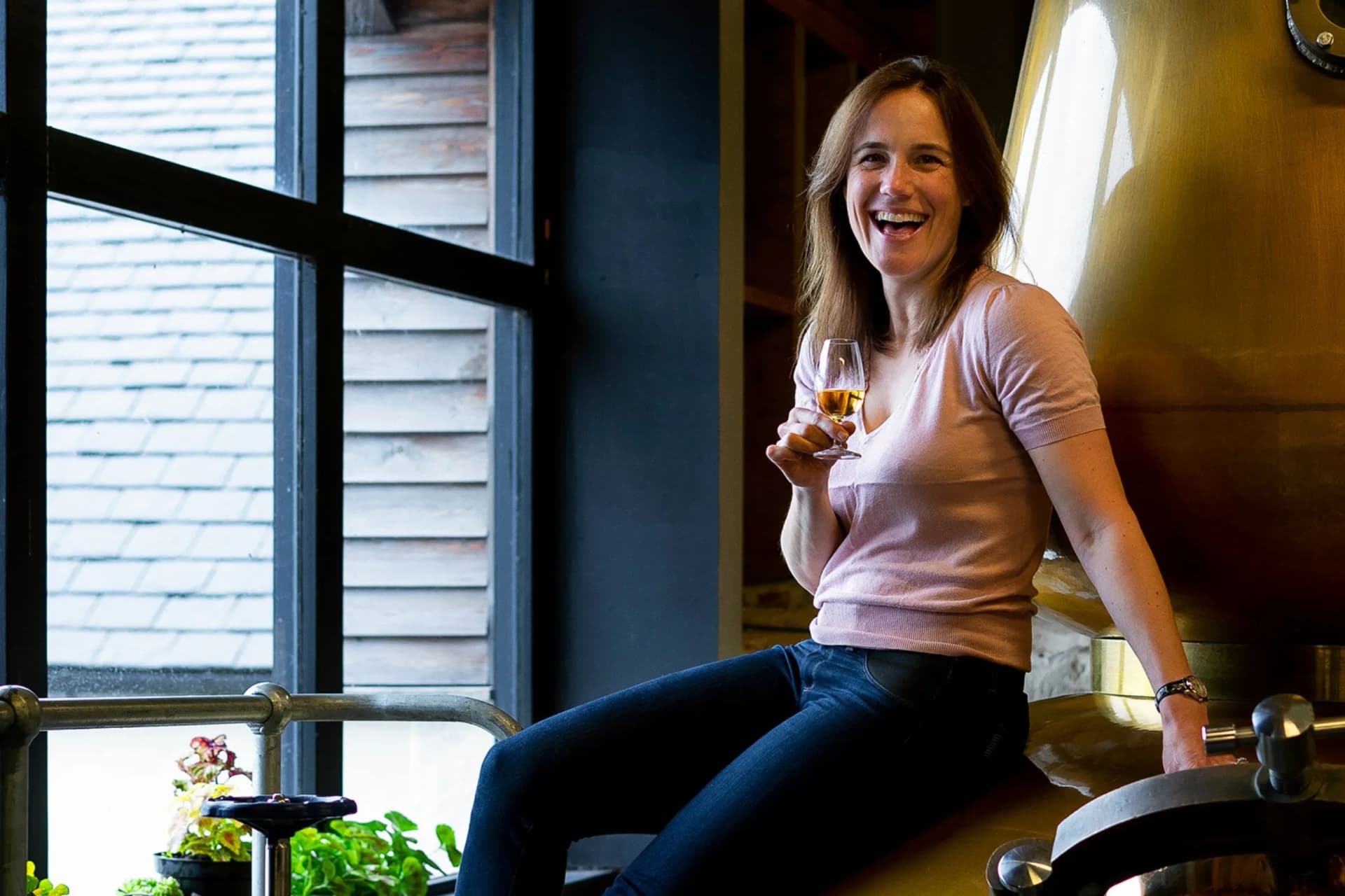 Smiling woman in a pink top and dark jeans sits on a metallic sculpture indoors, holding a glass of wine near a large window.