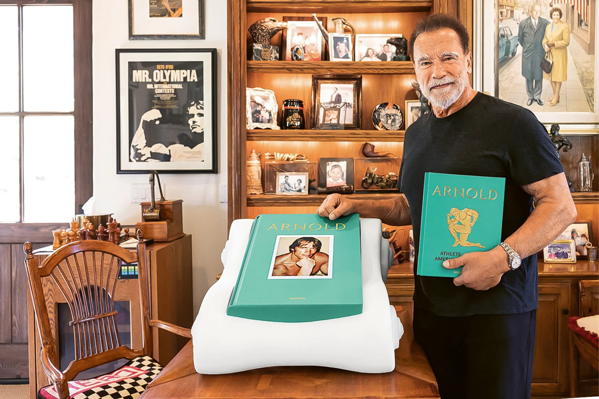 Older man with gray hair and beard in a black t-shirt stands in a room full of photos and memorabilia, holding a teal book and a teal pillow.