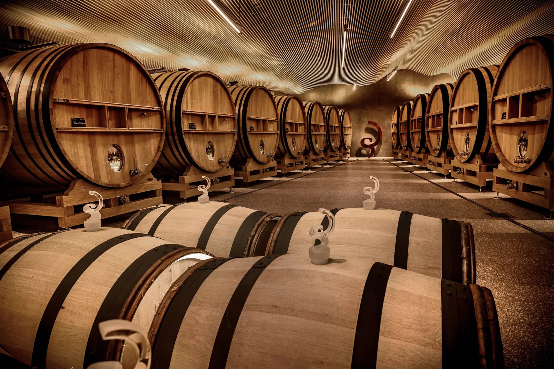 Warm winery cellar with rows of large wooden barrels lining shelves on both sides, receding toward the far end under amber lighting.
