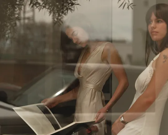 Two women in white dresses by a window, one looking down at a large book while the other faces forward; glass reflections create a soft, layered scene.