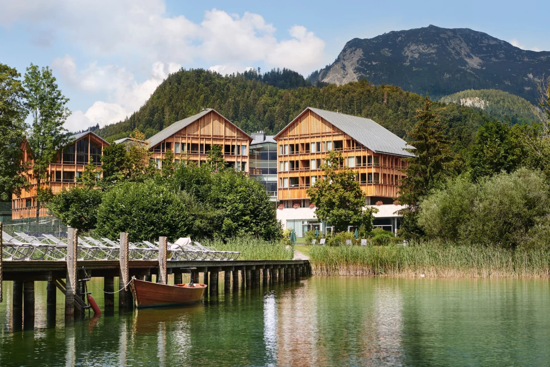 Chalet-style buildings sit beside a calm lake, with a wooden dock and small boat, surrounded by trees and distant mountains.