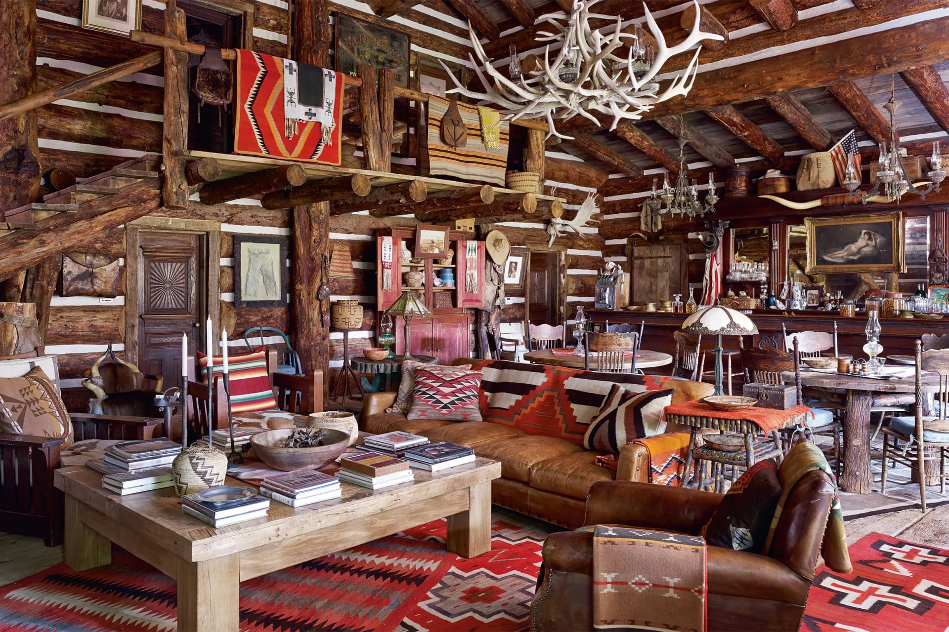 Rustic log cabin living room with antler chandelier, wooden beams, leather sofas, colorful rugs, woven textiles, and books on a coffee table.
