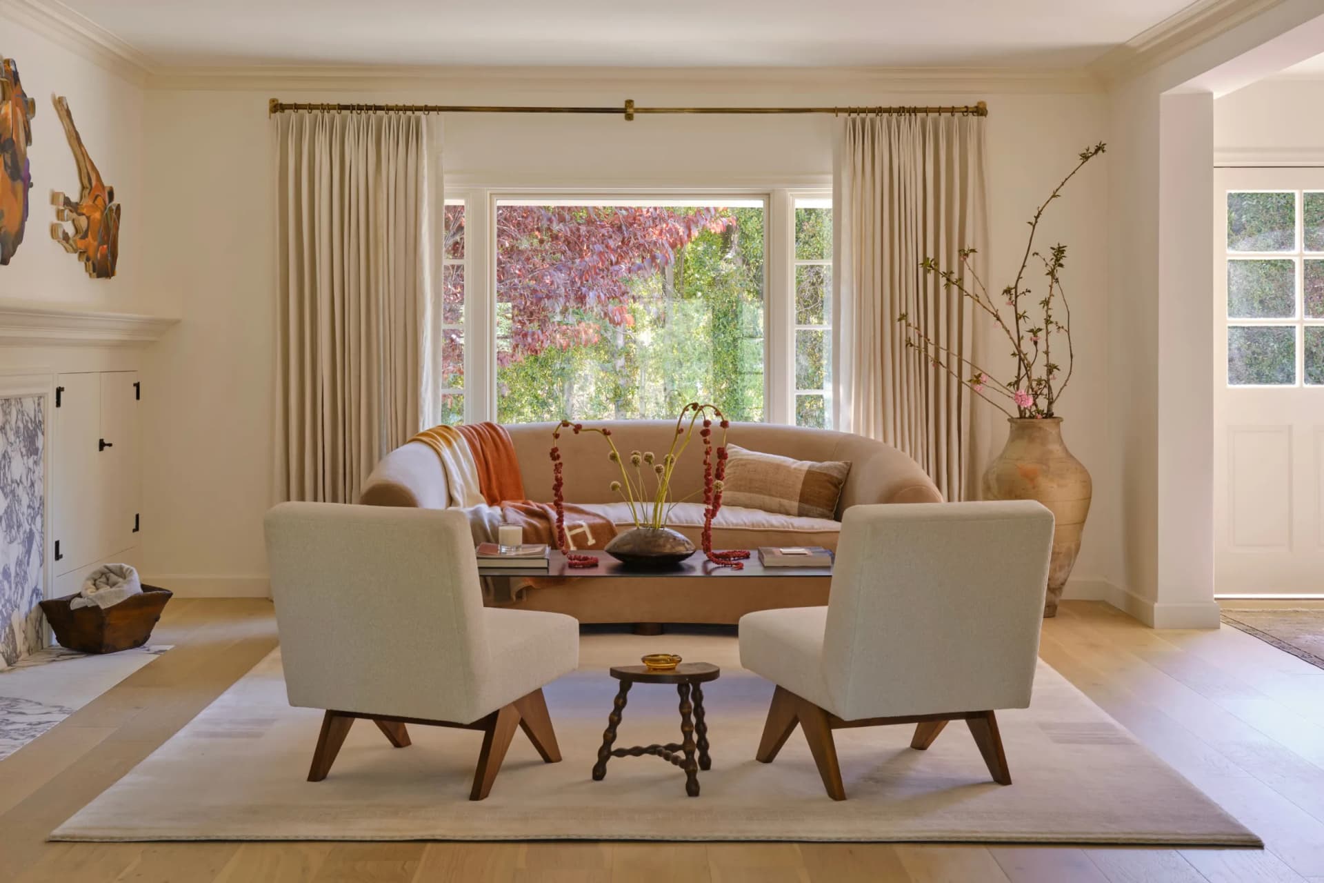 Bright living room with a cream sofa and two beige armchairs around a wooden coffee table on a light rug; vase with branches by the windows.
