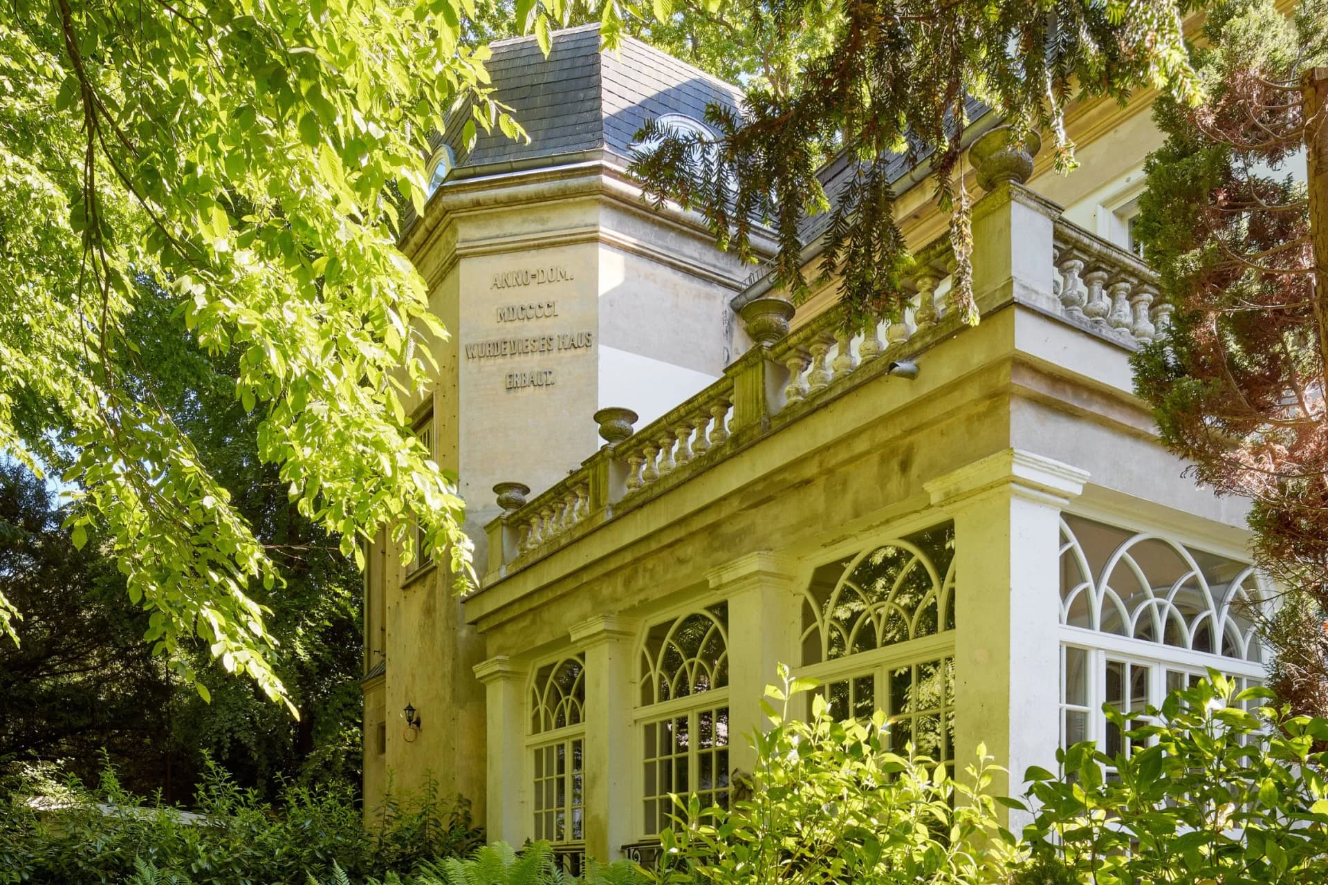 A sun-drenched historic villa with a balcony featuring a stone balustrade, arched windows and a slate roof, partly concealed by dense green foliage.