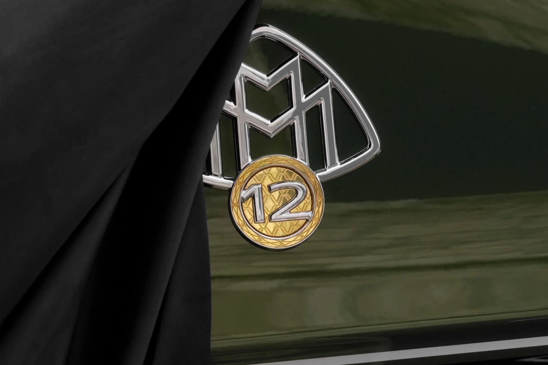A close-up of a chrome-plated emblem featuring an M-shaped motif, from which hangs a gold medal bearing the inscription ‘12’ against a dark background.