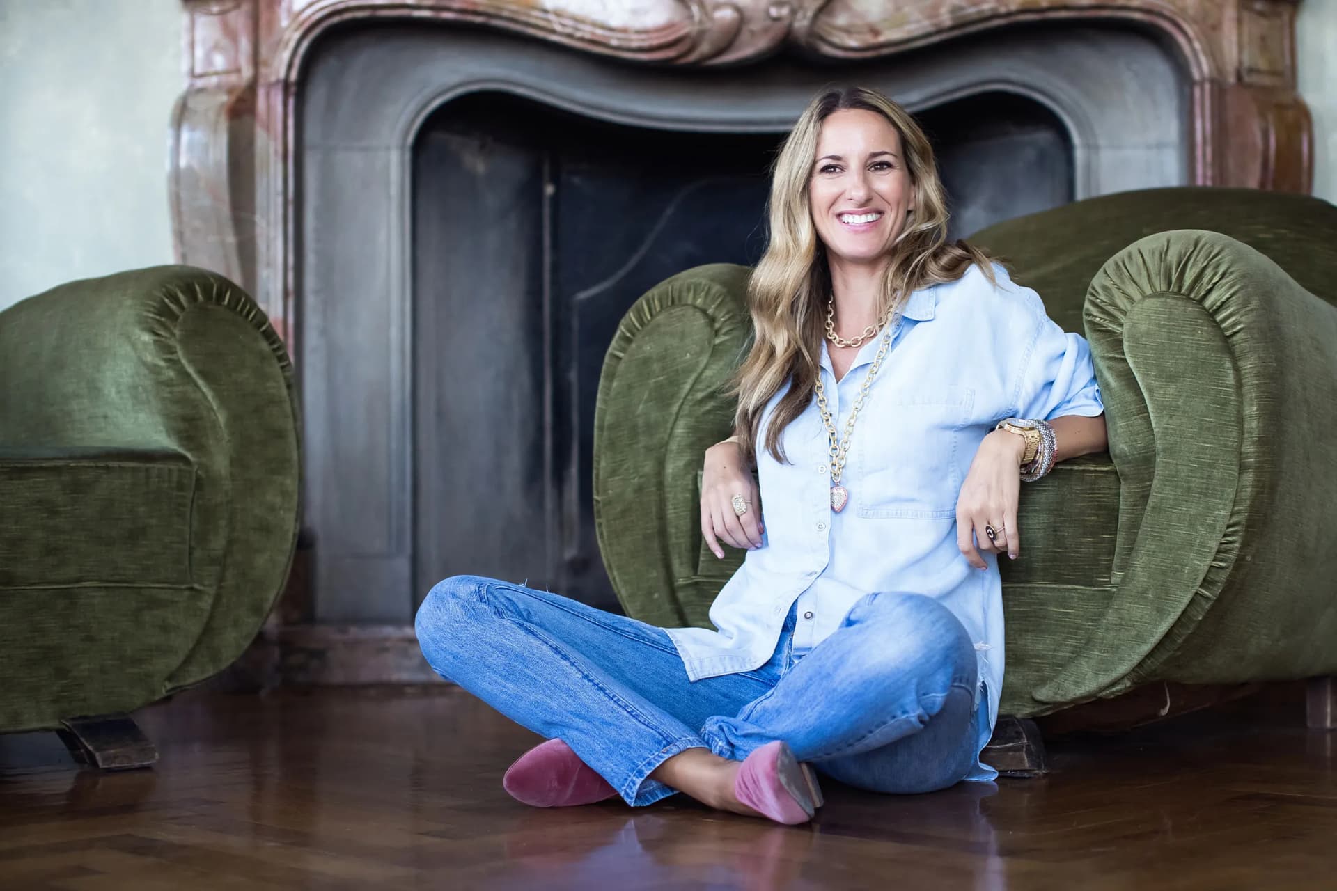 A smiling blonde woman in a light denim shirt and jeans is sitting cross-legged between two large green velvet chairs, in front of a vintage fireplace.