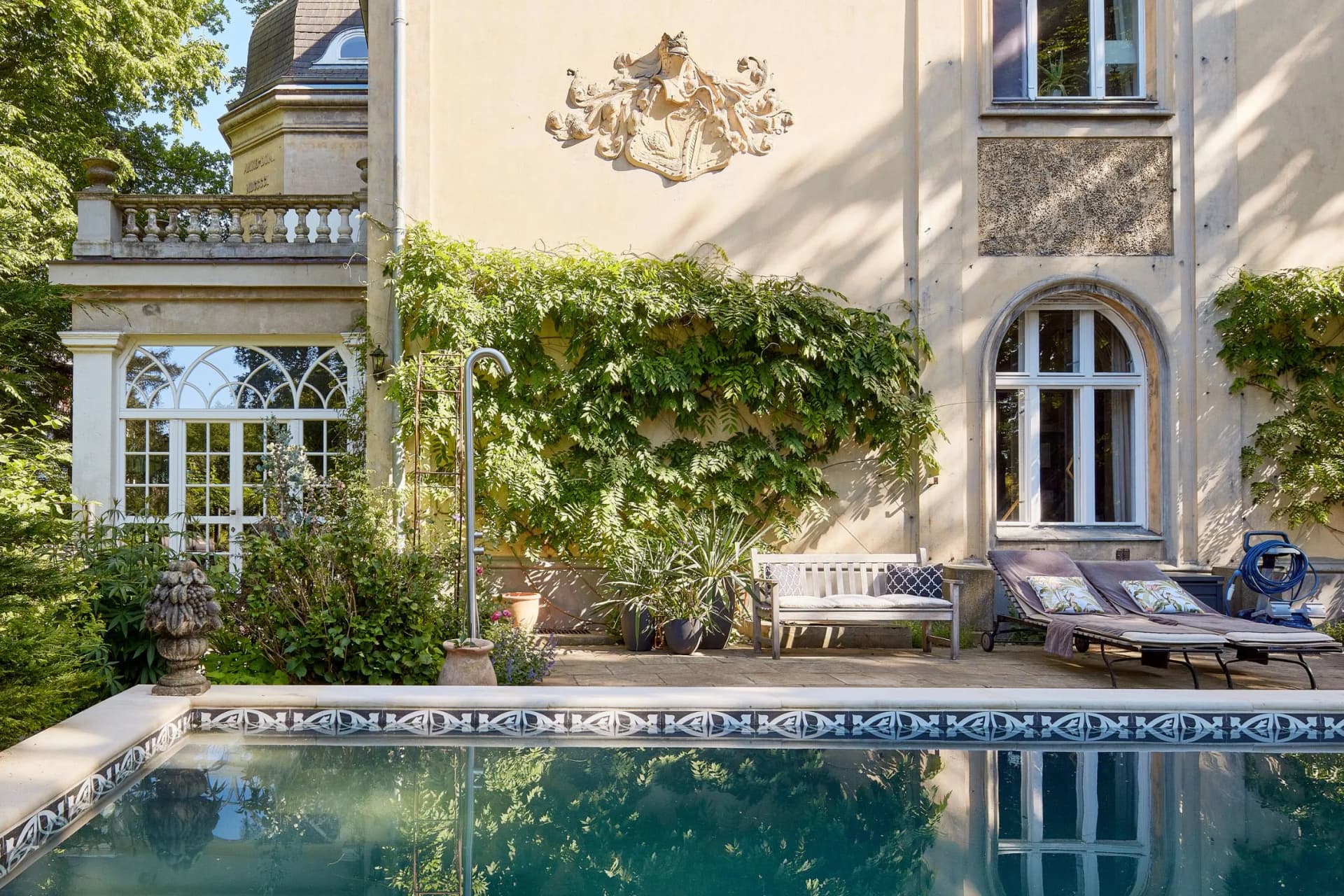A sun-drenched beige villa with an ornate coat of arms on the wall, an ivy-covered façade, a balcony with a balustrade on the left-hand side, an arched window and a reflecting pool with sun loungers.