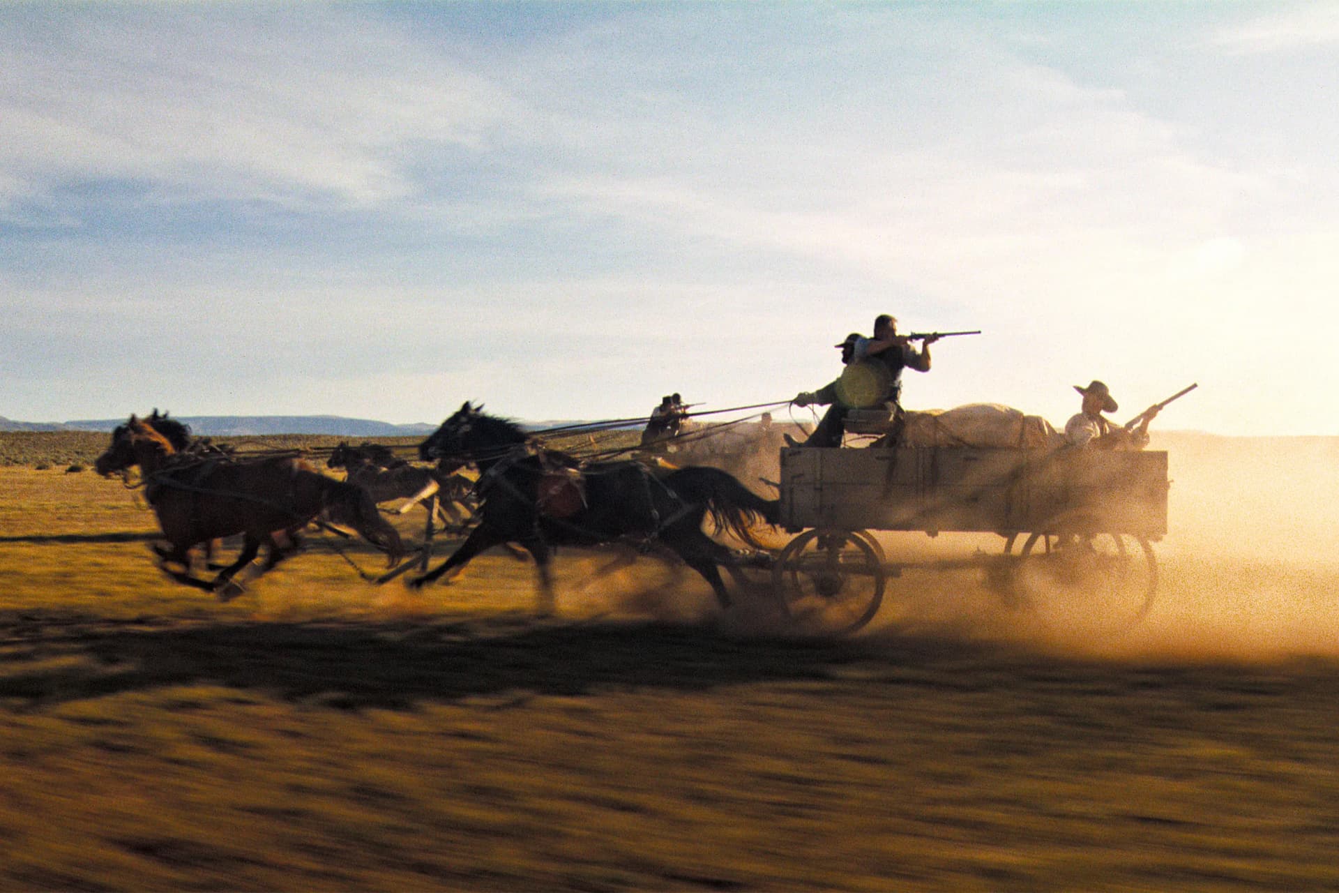 Dusty desert plain at sunset with a team of horses pulling a wooden wagon, riders on and around the wagon as it kicks up dust.