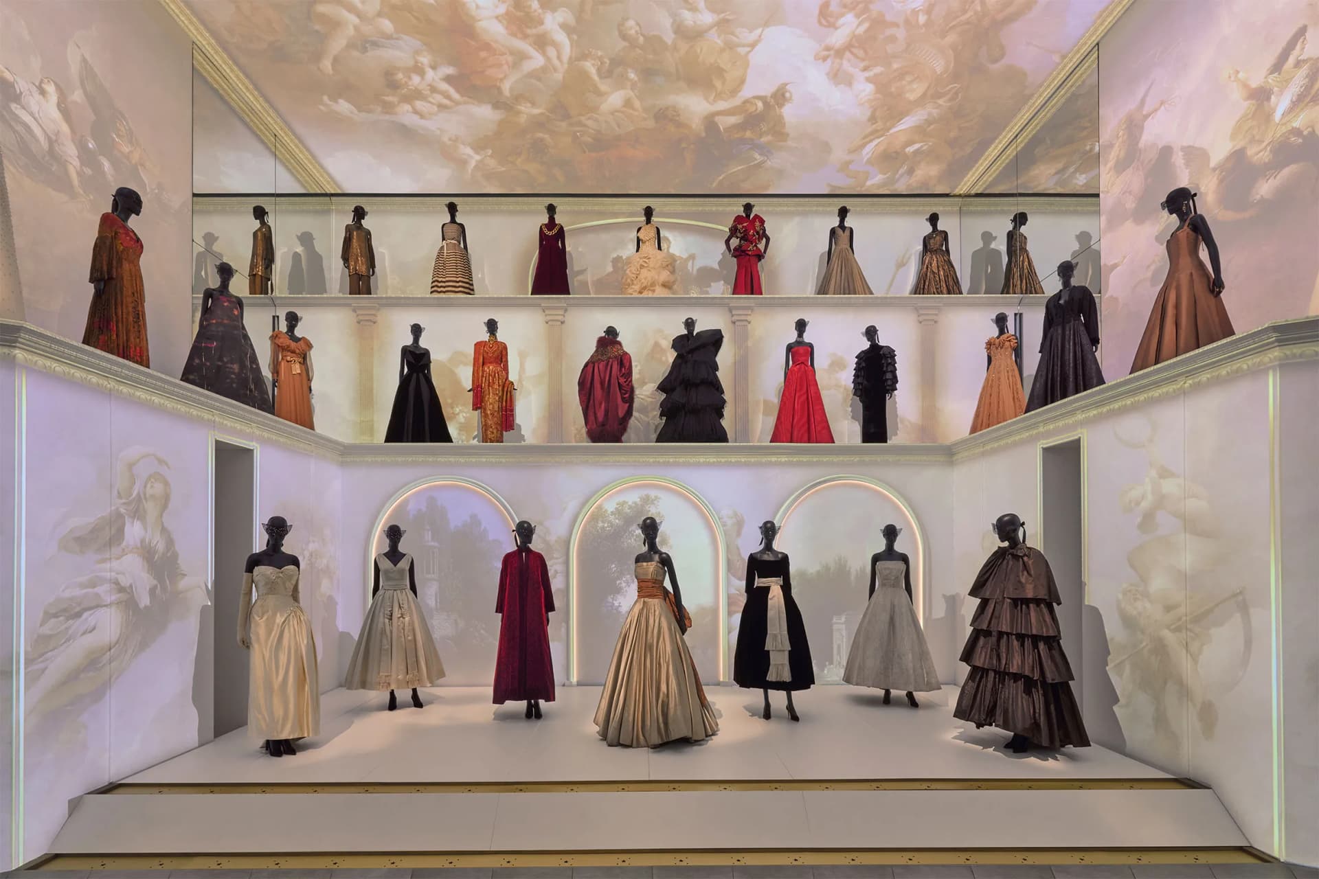 Two-tier display of elegant gowns on black mannequins in a grand museum, with a painted ceiling mural overhead.