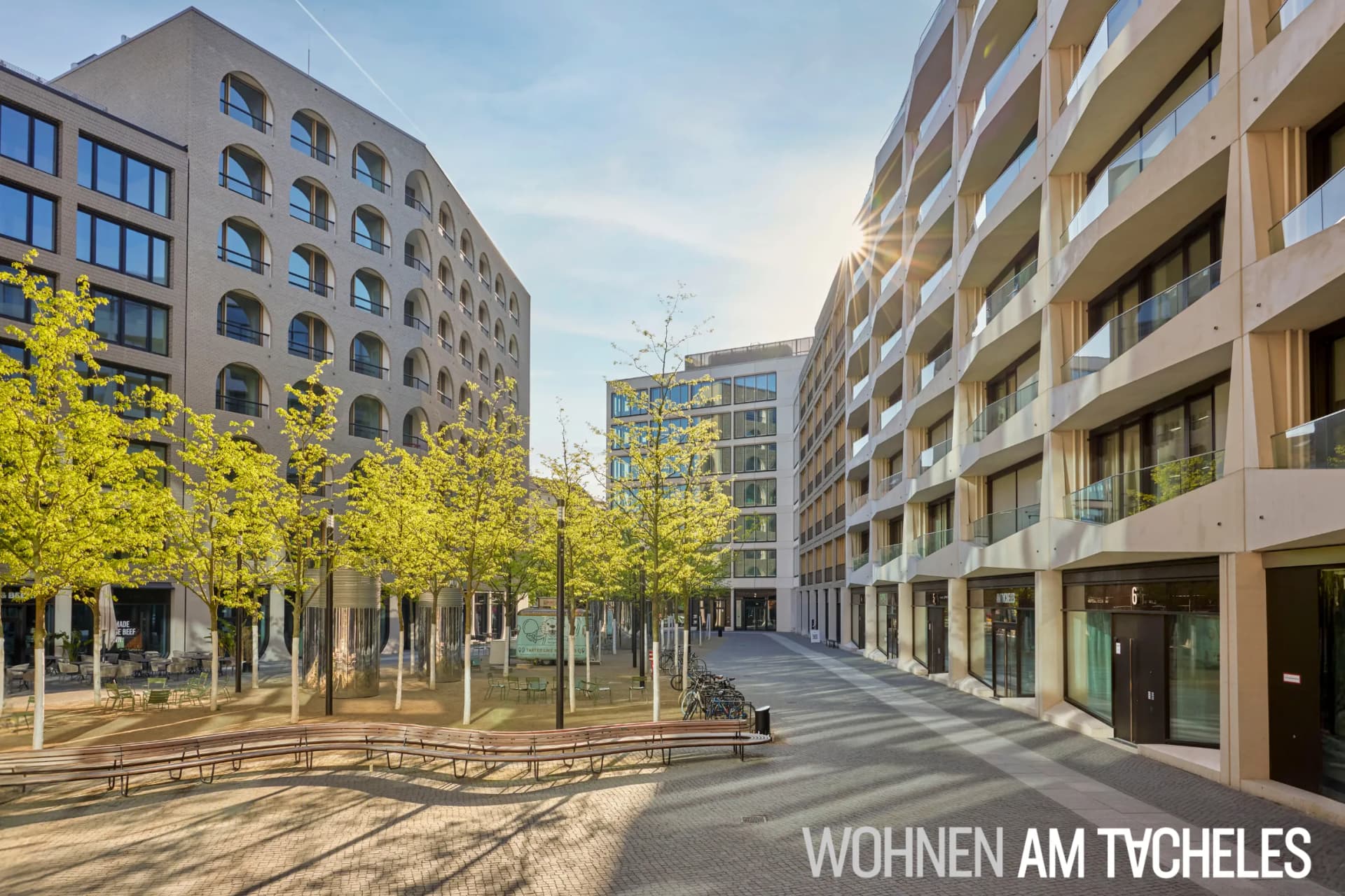 Sunlit plaza between a beige brick building with arched windows and a modern white building with glass balconies, lined by trees.