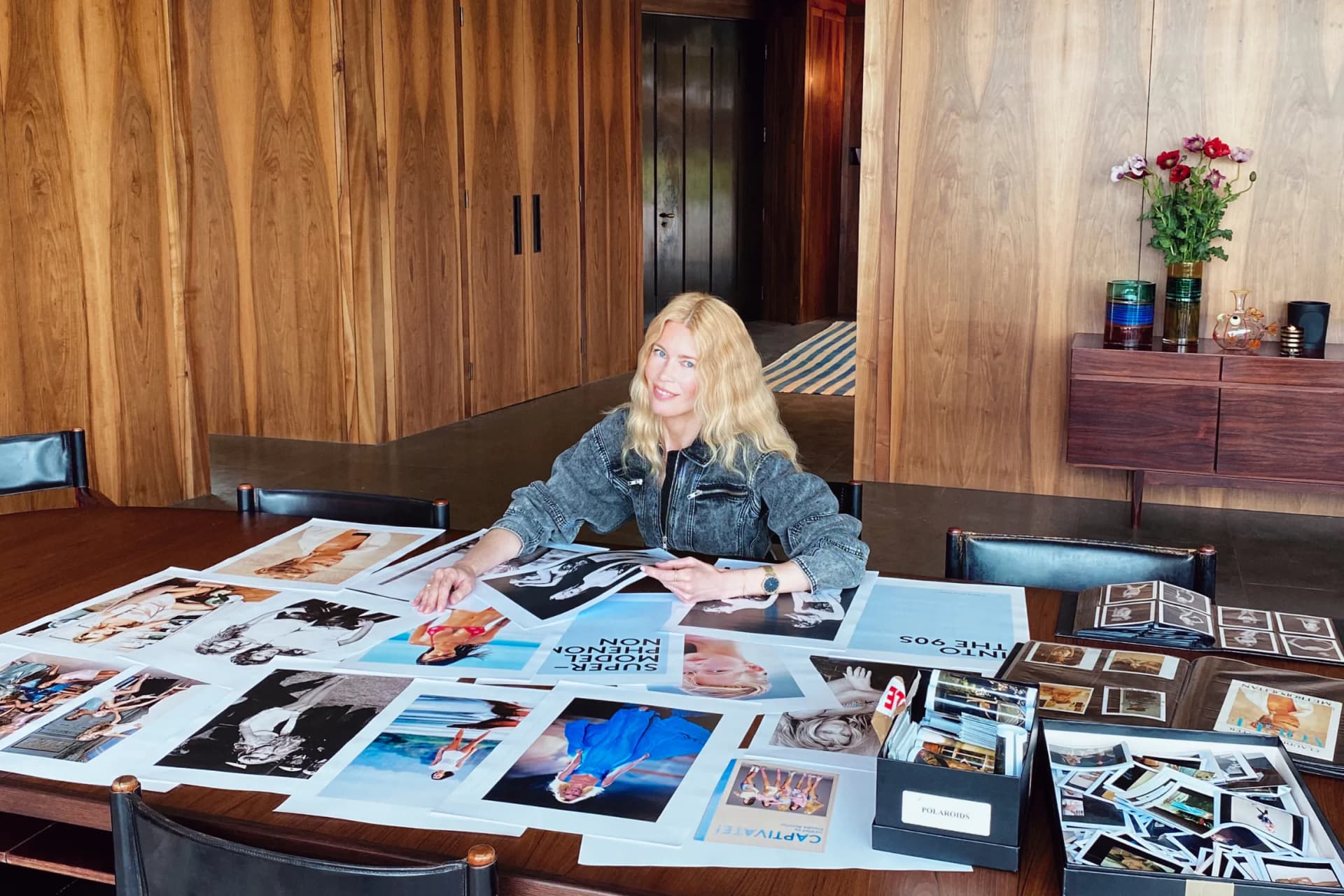 Smiling blonde woman sits at a long wooden table covered with numerous photos, in a wood-paneled room with a sideboard and flowers.